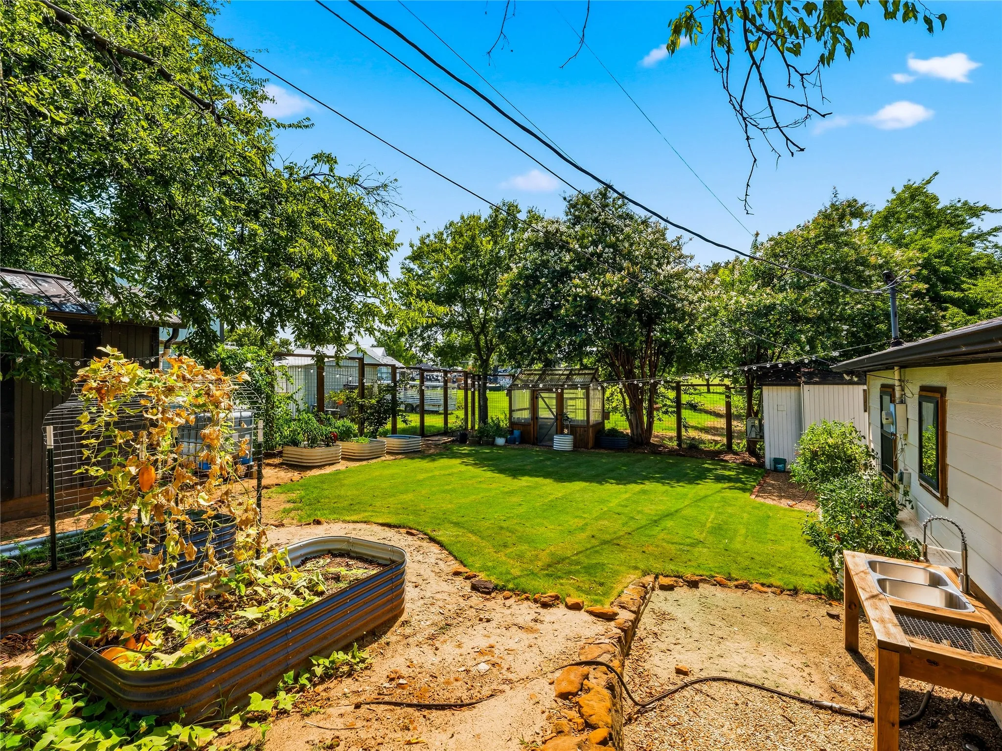 Expansive backyard featuring a manicured lawn, multiple raised garden beds, a greenhouse, and mature trees