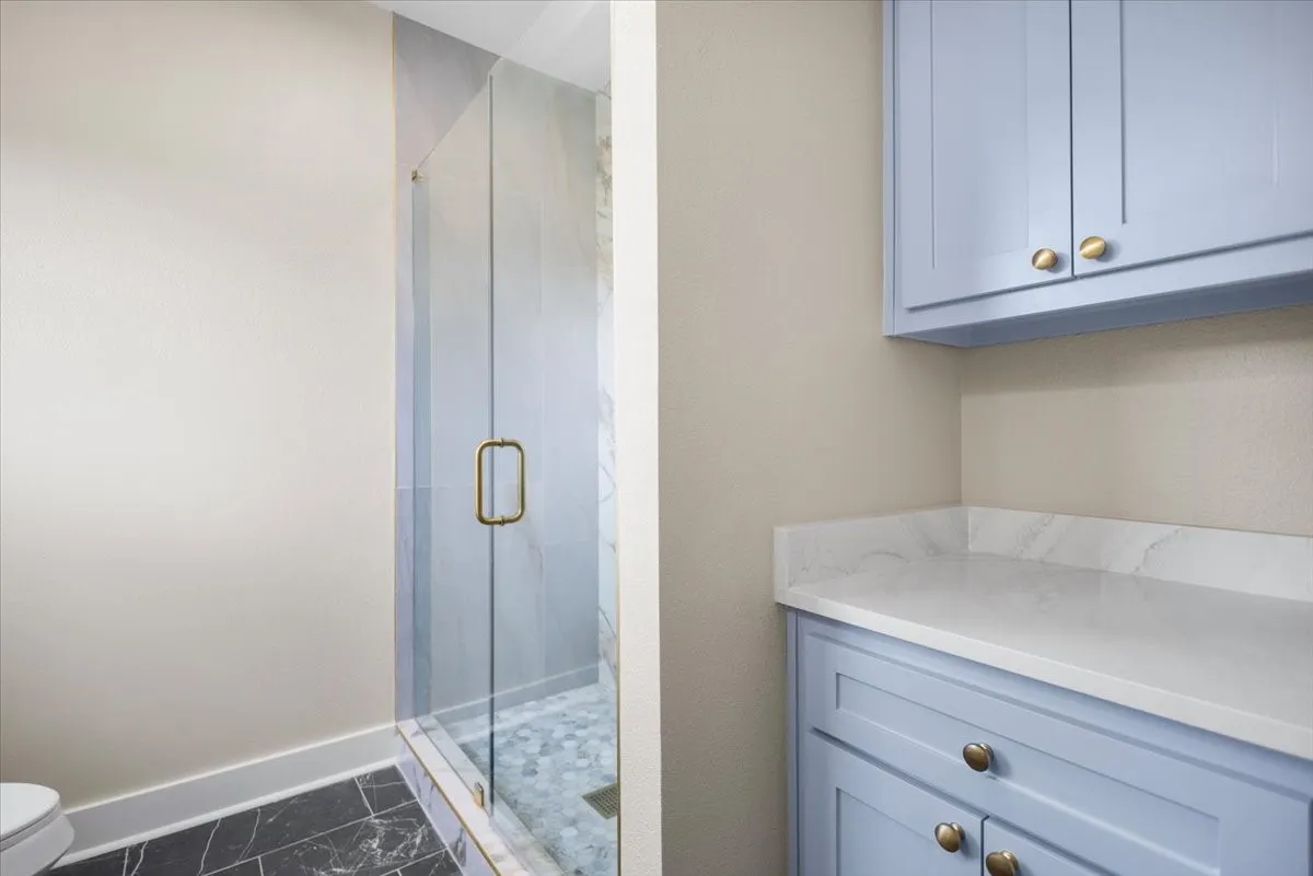 Glass-enclosed shower with gold-tone hardware, marble-look wall tiles, and pebble-style floor tiles