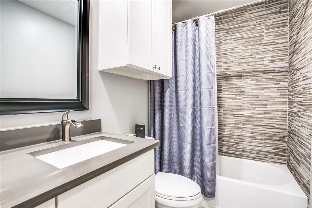 Bathroom featuring a built-in tub with textured tile surround, a white vanity with integrated sink and gray countertop, brushed nickel faucet, and overhead cabinetry