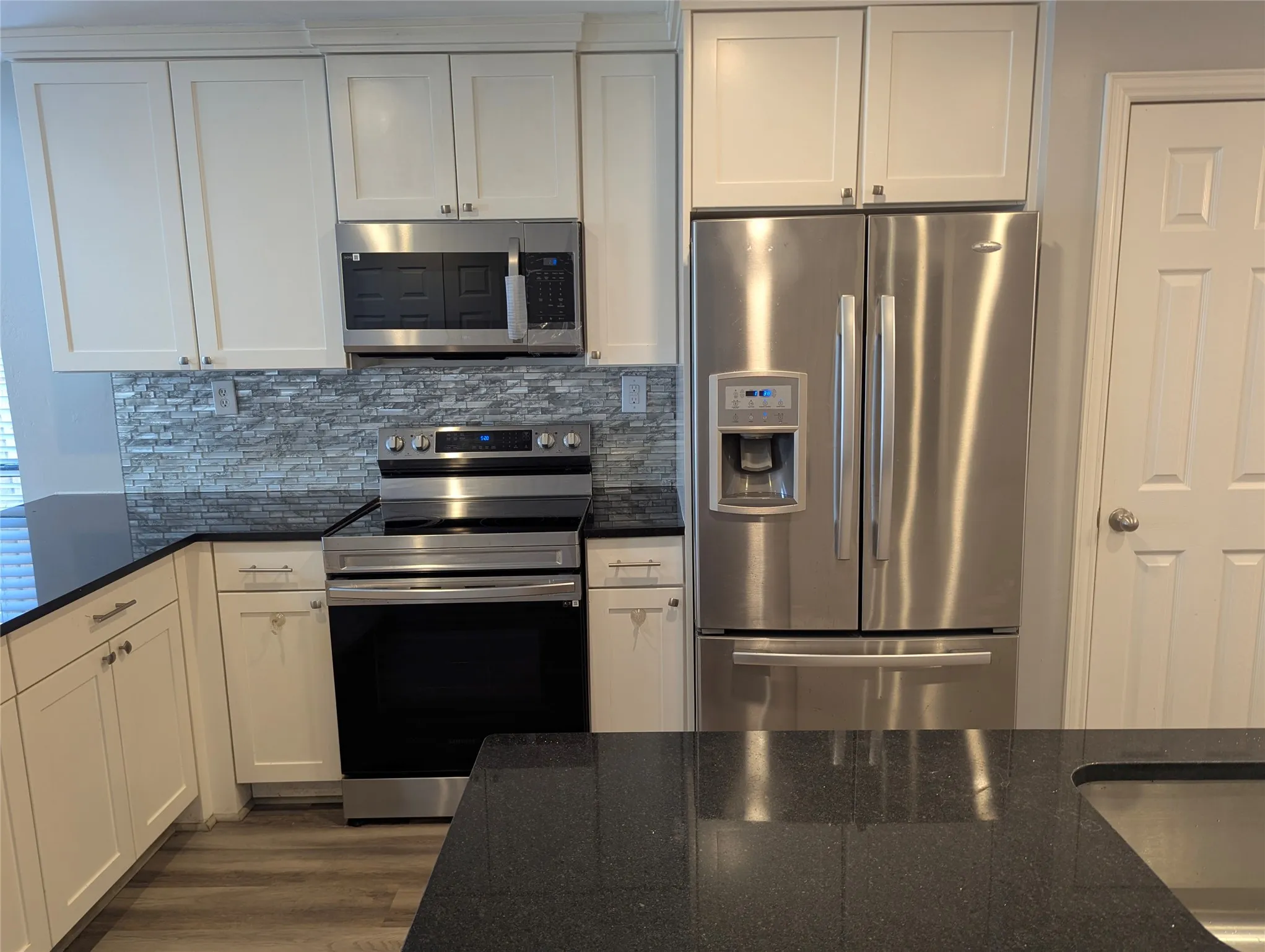 White shaker cabinetry, dark countertops, full suite of stainless steel appliances, stacked stone backsplash, and wood-finish flooring