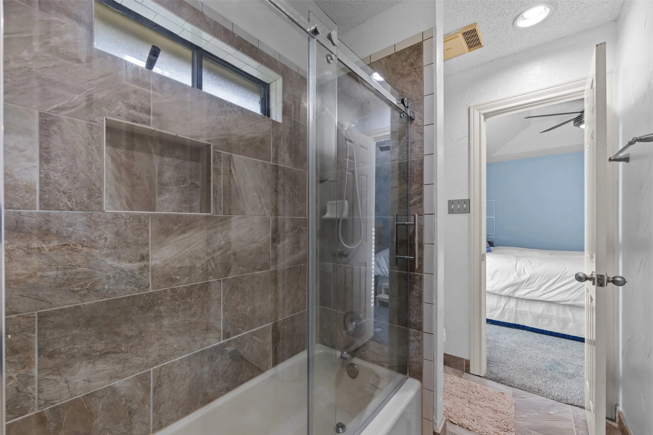 Bathtub with a glass sliding shower door featuring large format stone-look tile surround, a built-in shower niche, and a transom window