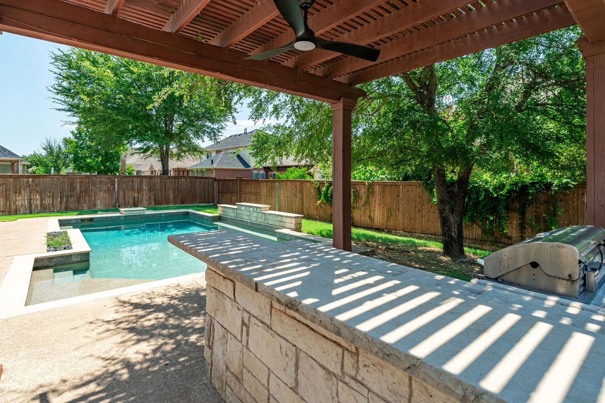 Outdoor entertainment area featuring a custom pool, stone-clad outdoor kitchen, and a wooden pergola with a ceiling fan