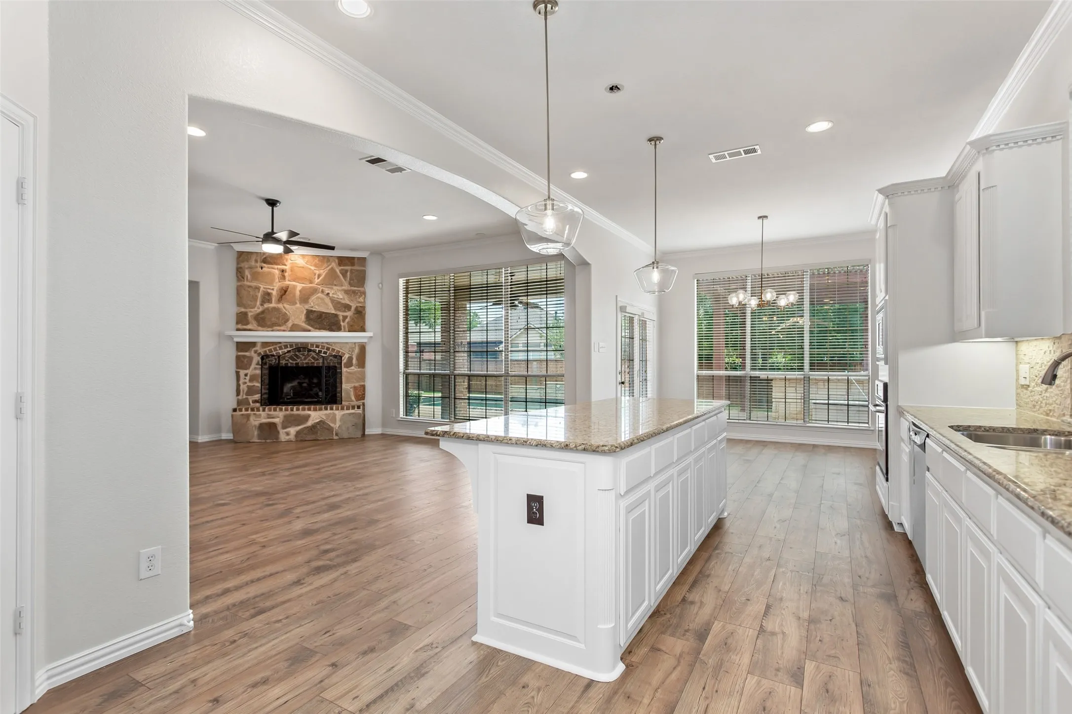 Open-concept interior featuring light wood-finish flooring, a kitchen island with granite countertop, white paneled cabinetry, and a stone fireplace