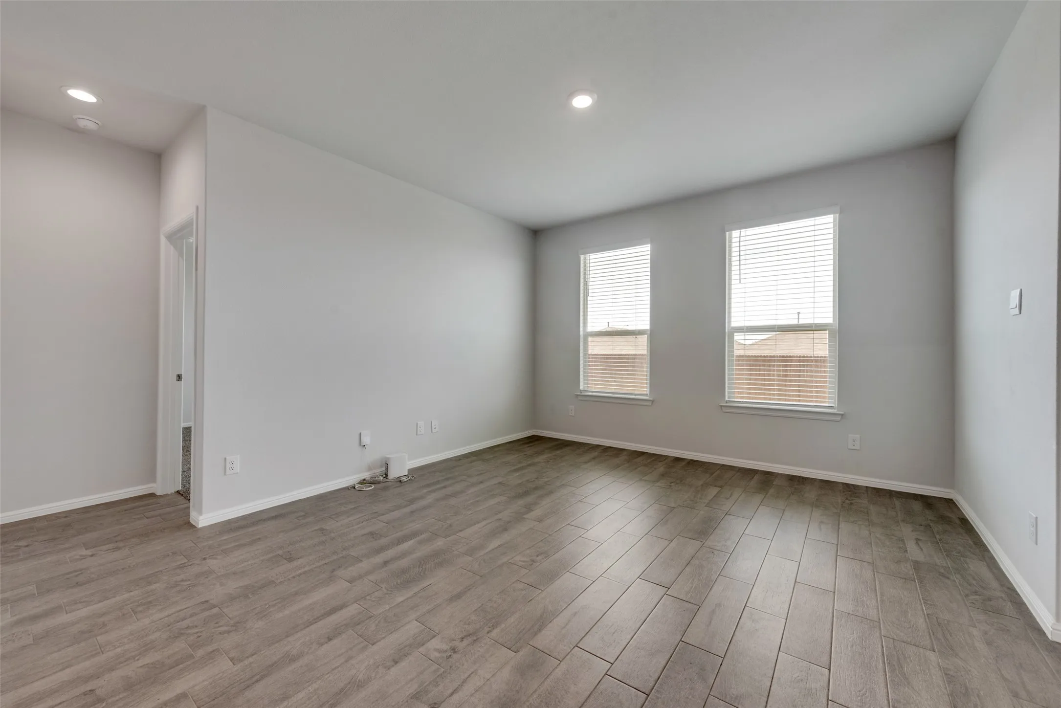 Spacious interior room featuring wood-finish plank flooring, recessed ceiling lighting, and two windows with blinds