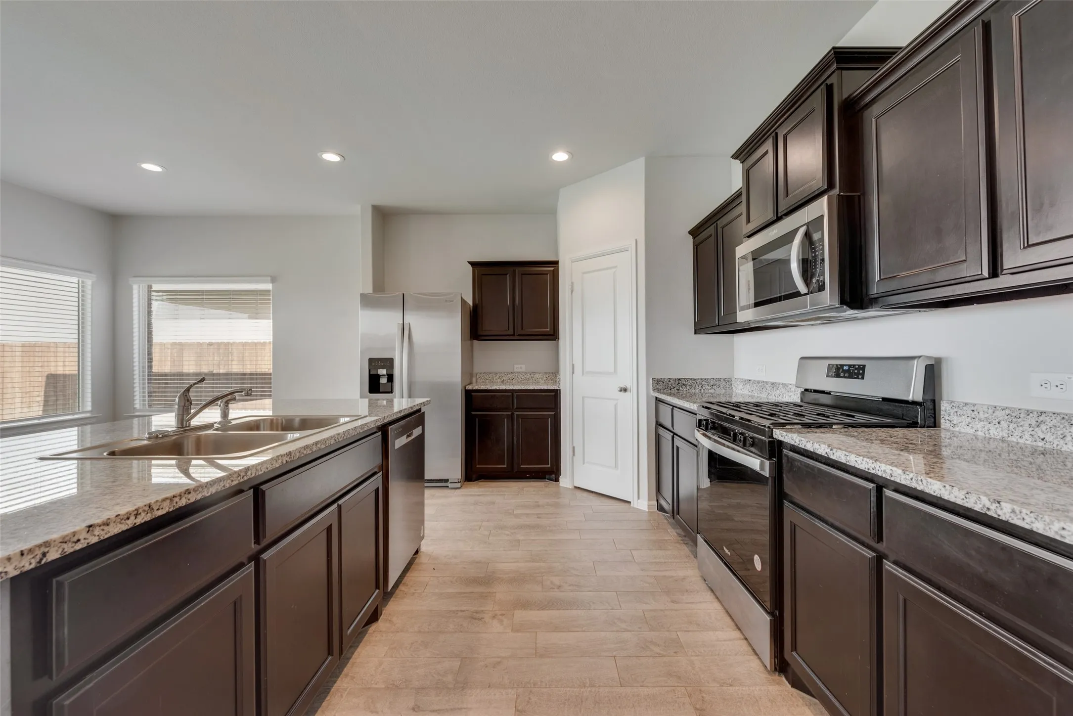 Kitchen featuring dark wood cabinetry, light granite countertops, stainless steel appliances, a kitchen island with an undermount sink, and wood-finish flooring