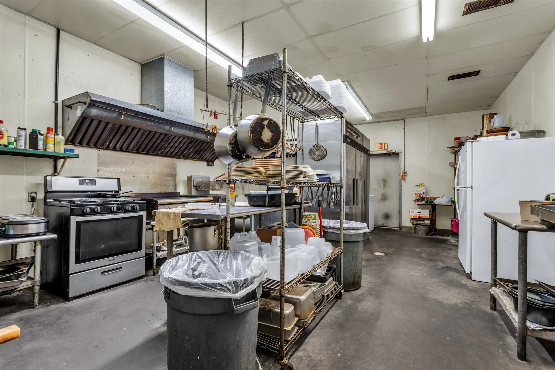 Commercial kitchen featuring a stainless steel range with overhead ventilation hood, industrial shelving, and a large refrigerator