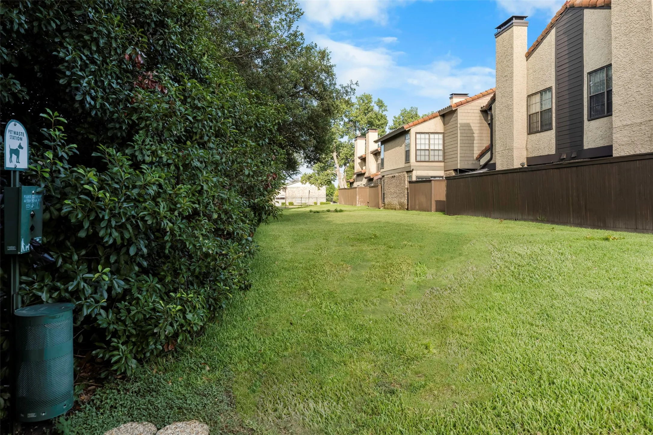 Expansive green space featuring a pet waste station with a dispenser and receptacle, bordered by a substantial privacy hedge and a wooden fence