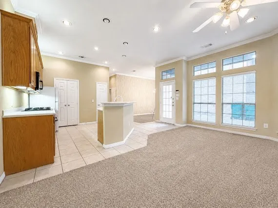 Open-concept living area featuring a kitchen with wood cabinetry and tile flooring, a living space with carpet, a ceiling fan, and large windows, and a door with a glass insert