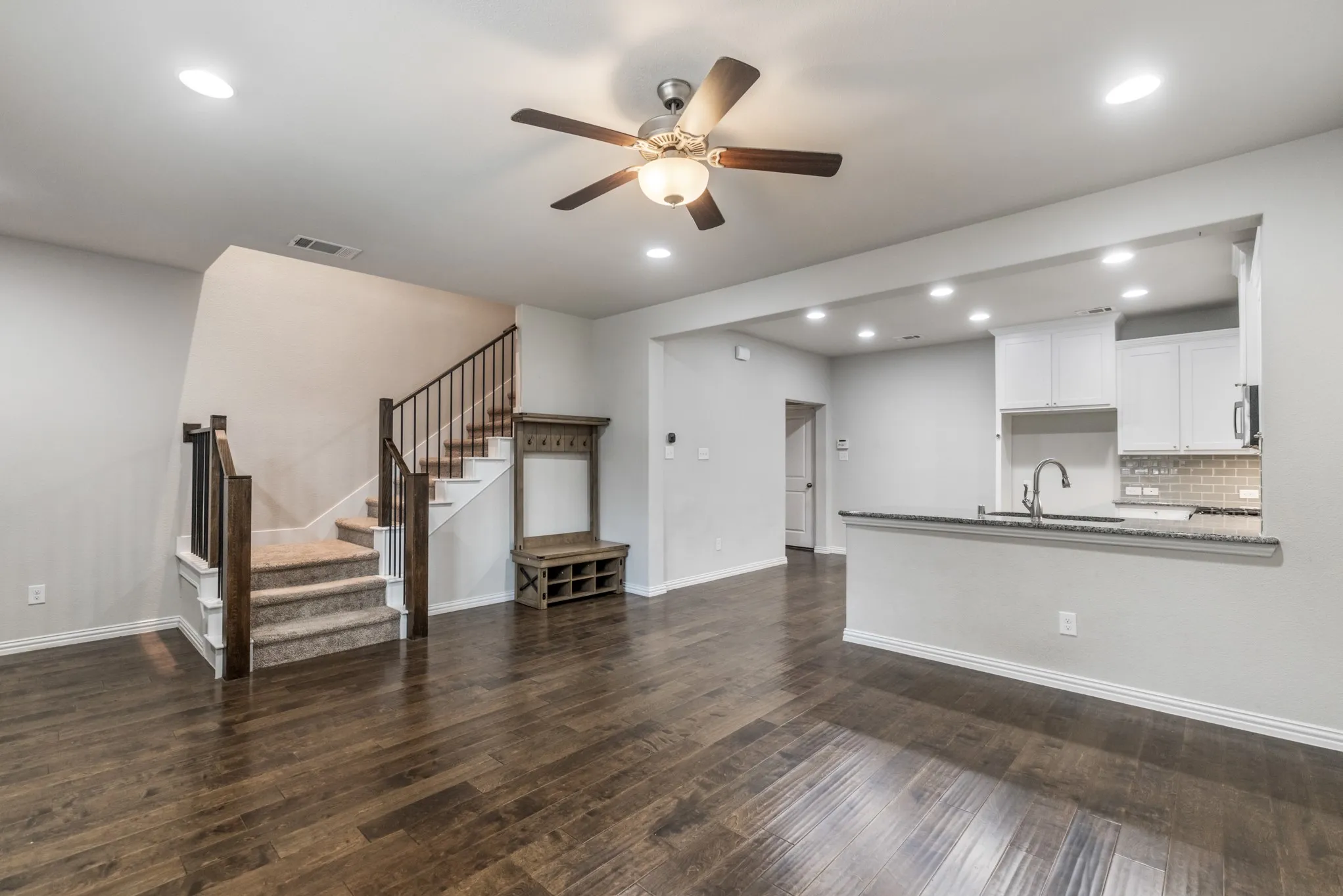 Spacious interior featuring wood-finish flooring, a contemporary staircase with carpeted treads and dark wood newel posts, a kitchen with white cabinetry and a breakfast bar, recessed lighting, and a ceiling fan with integrated lighting