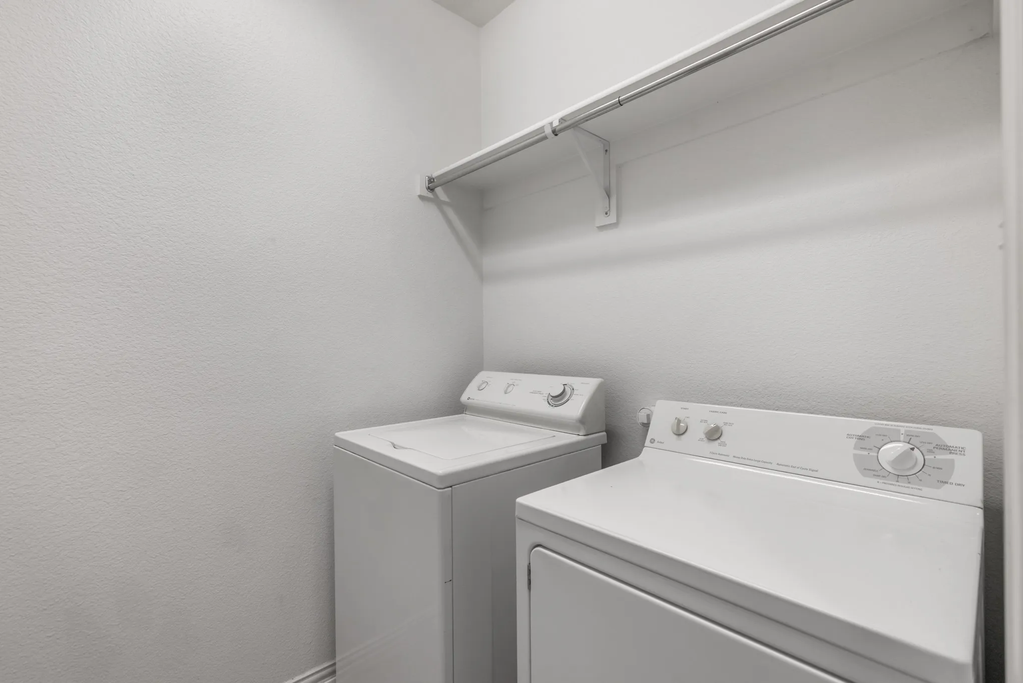 Dedicated laundry area featuring white textured walls, a white top-load washing machine, a white front-load dryer, and an overhead storage shelf