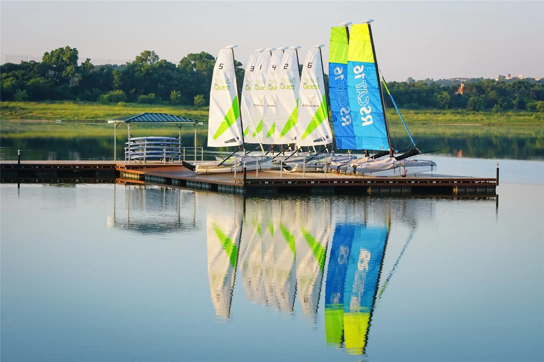 Floating dock with multiple sailboats, a covered paddleboard storage area, and tranquil water reflections