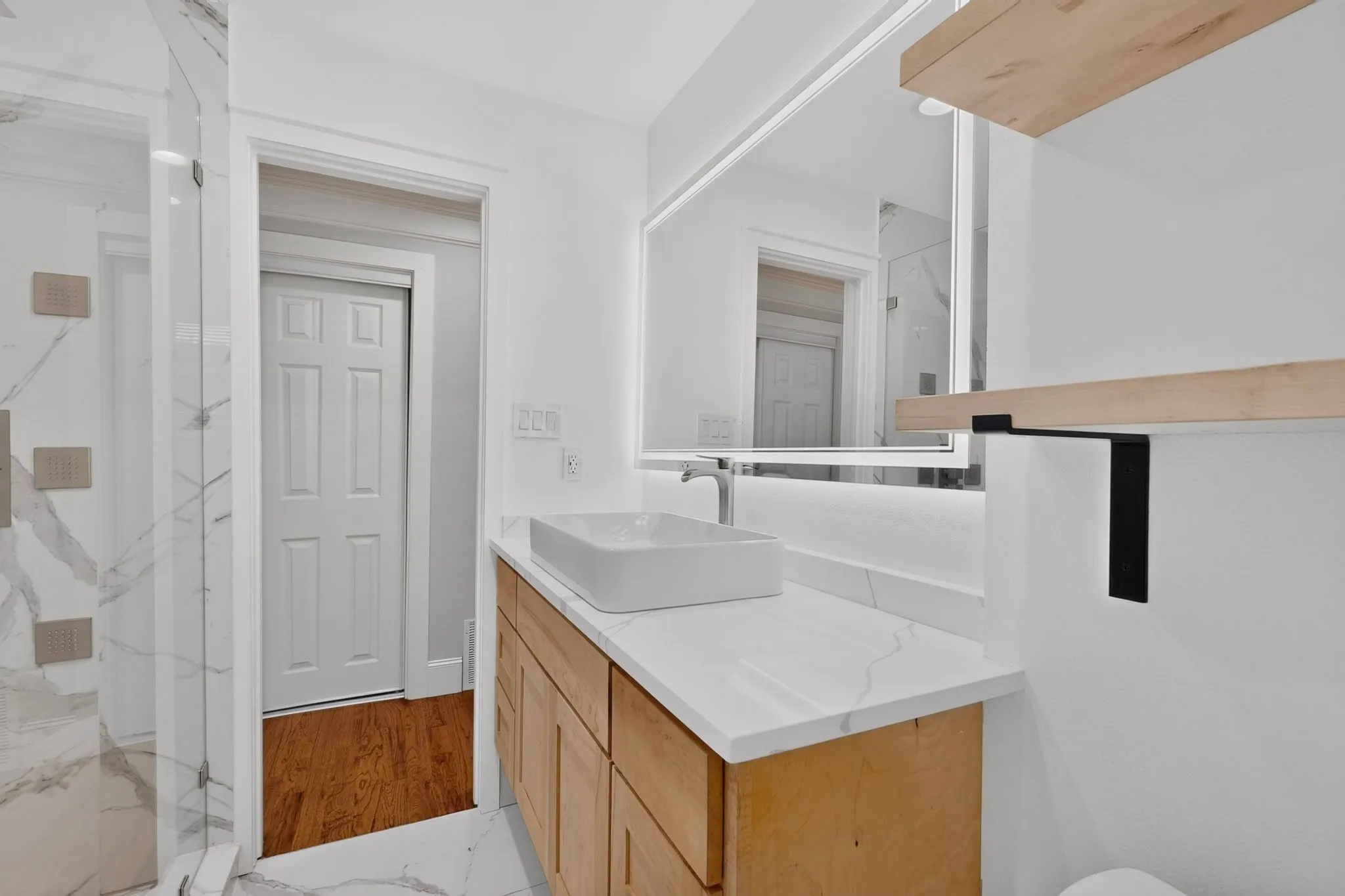 Modern bathroom featuring a light wood-finish vanity with a white vessel sink, a white countertop with subtle veining, and an illuminated mirror