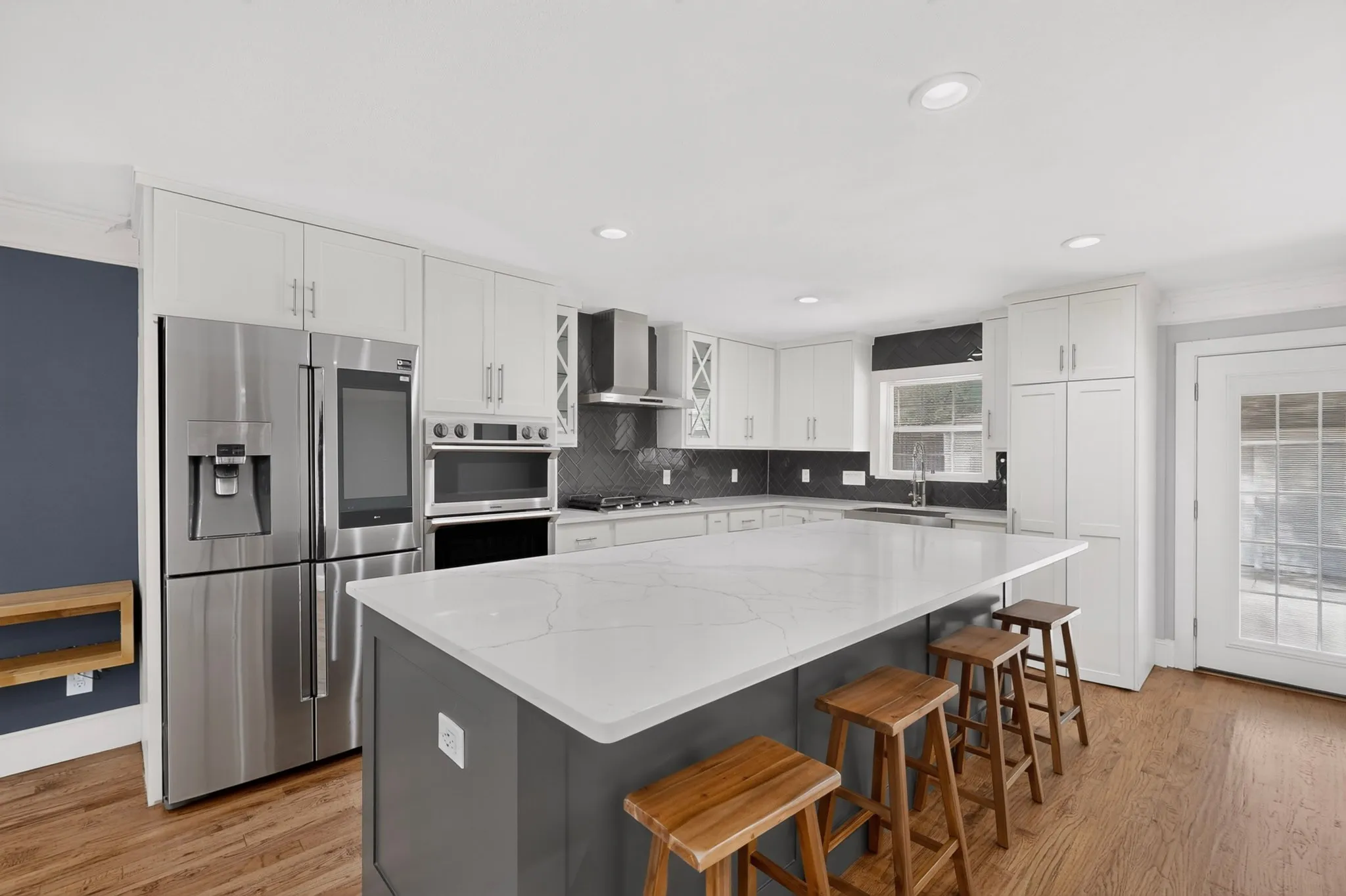 Gourmet kitchen with a large central island featuring a light-toned countertop, stainless steel appliances, white cabinetry, dark backsplash, and wood-finish flooring