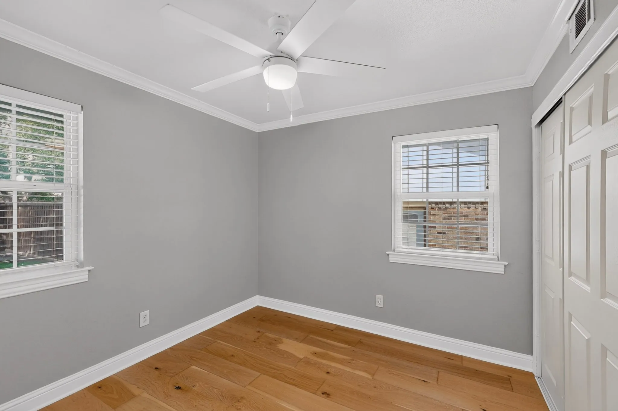 Interior room featuring wood-finish flooring, two windows with blinds, a ceiling fan with integrated lighting, crown molding, and a sliding closet door