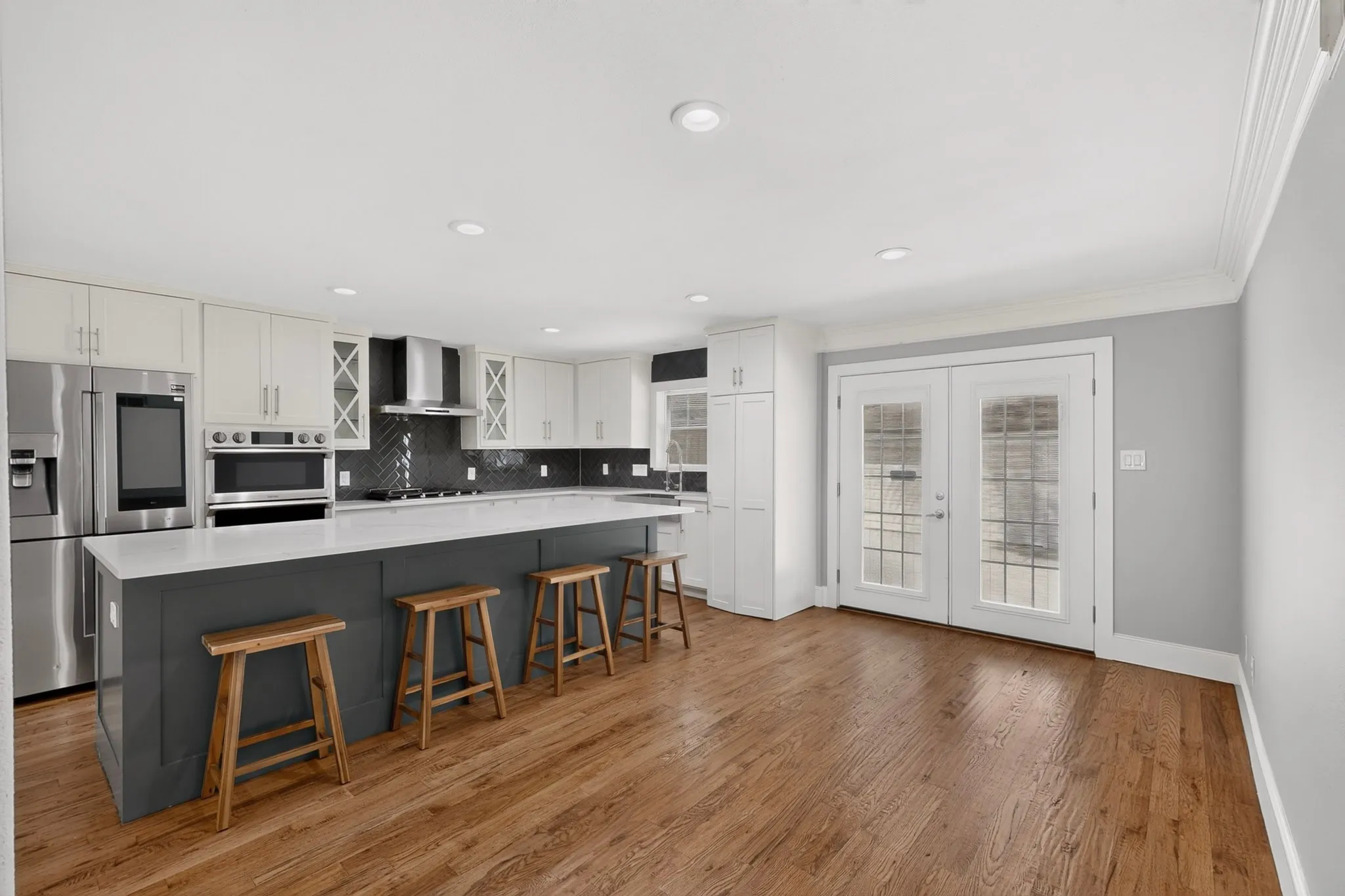 Contemporary kitchen featuring an oversized island, stainless steel appliances, herringbone tile backsplash, hardwood flooring, and French doors