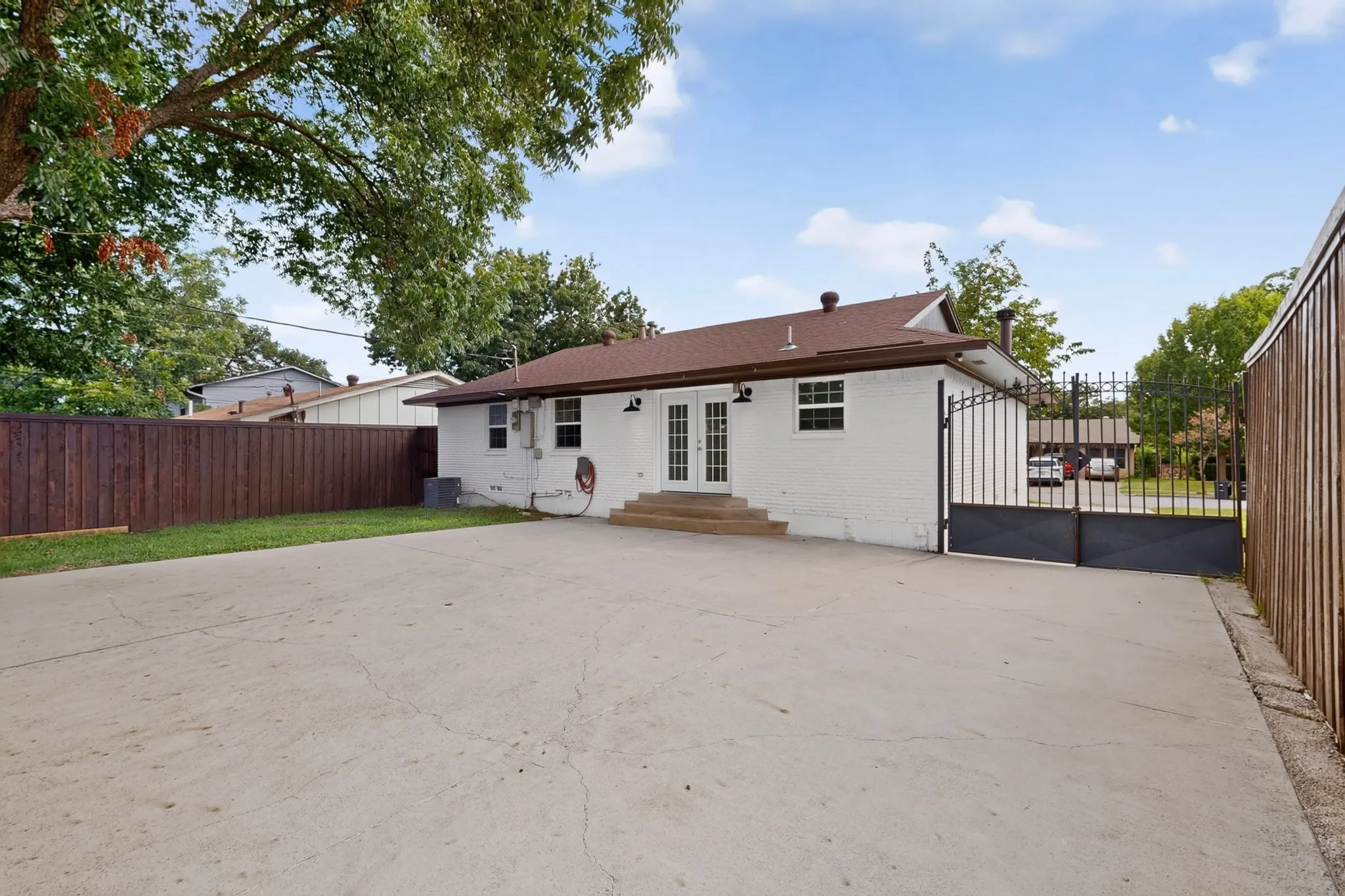 Expansive concrete patio with white brick exterior, double French doors, a dark wood fence, and a wrought iron gate