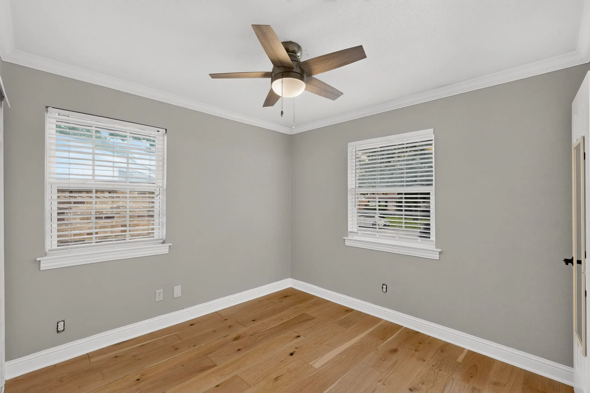 Room featuring wood-finish flooring, two windows with white blinds, a ceiling fan with integrated lighting, crown molding, and a solid door with a black handle