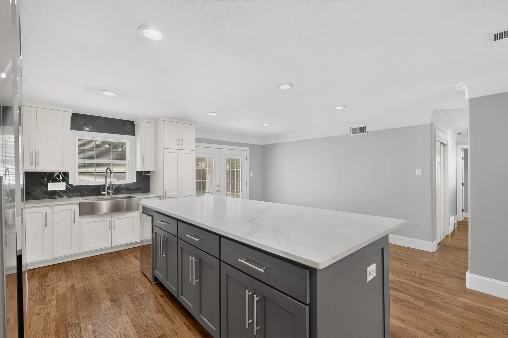 Contemporary kitchen featuring a large central island with a solid surface countertop, white upper and lower cabinetry, a dark herringbone pattern backsplash, and wood-finish flooring