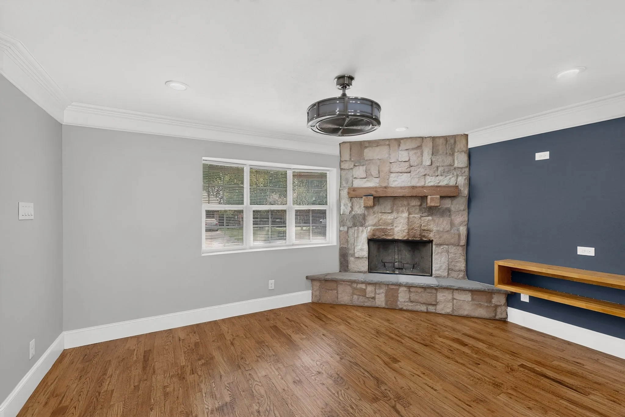Living area featuring a stone fireplace with a wood mantle, wood-finish flooring, a multi-pane window, and a built-in wood shelf