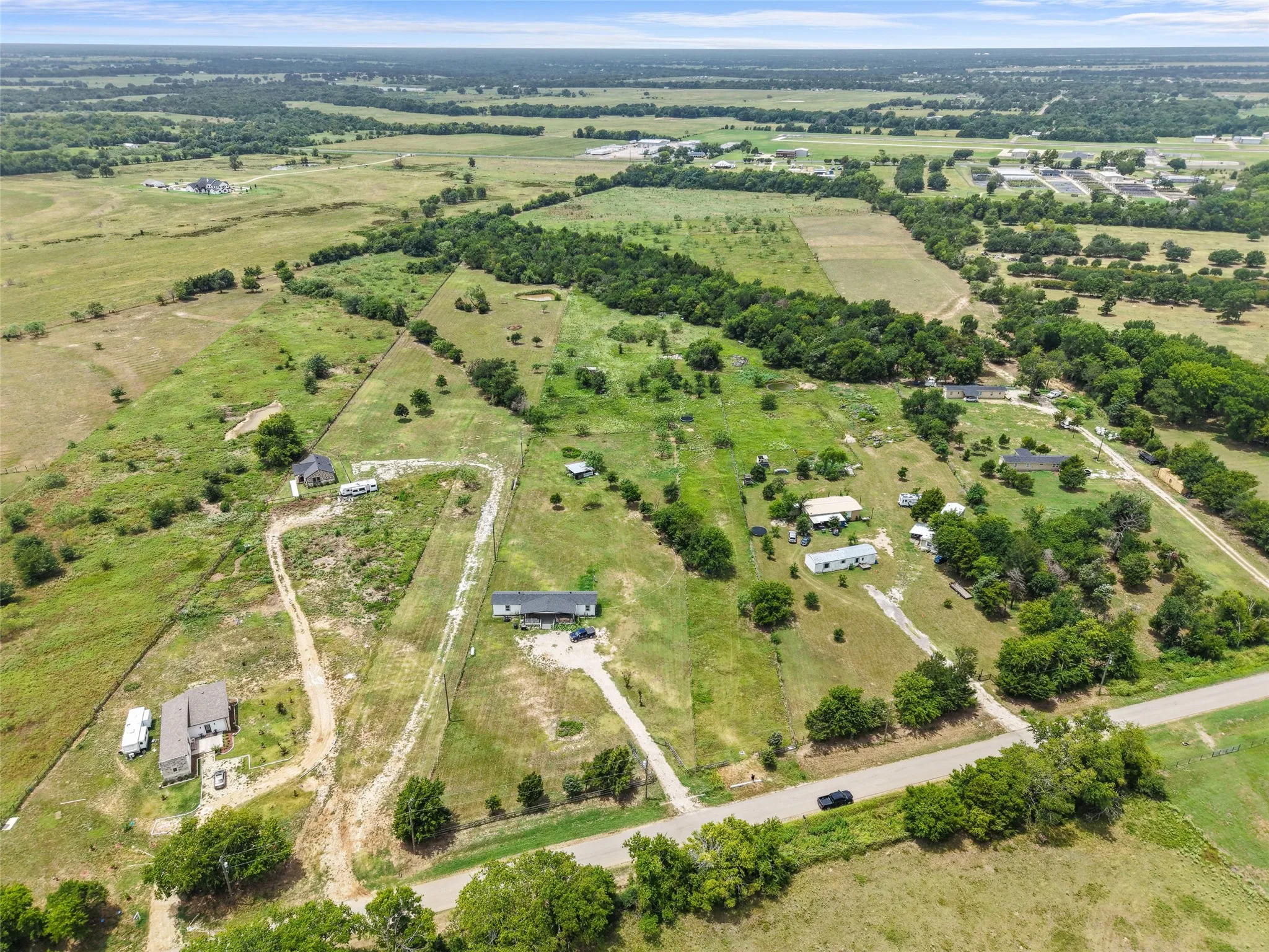 Expansive rural landscape featuring multiple structures, mature trees, and unpaved access paths, bordered by a paved roadway