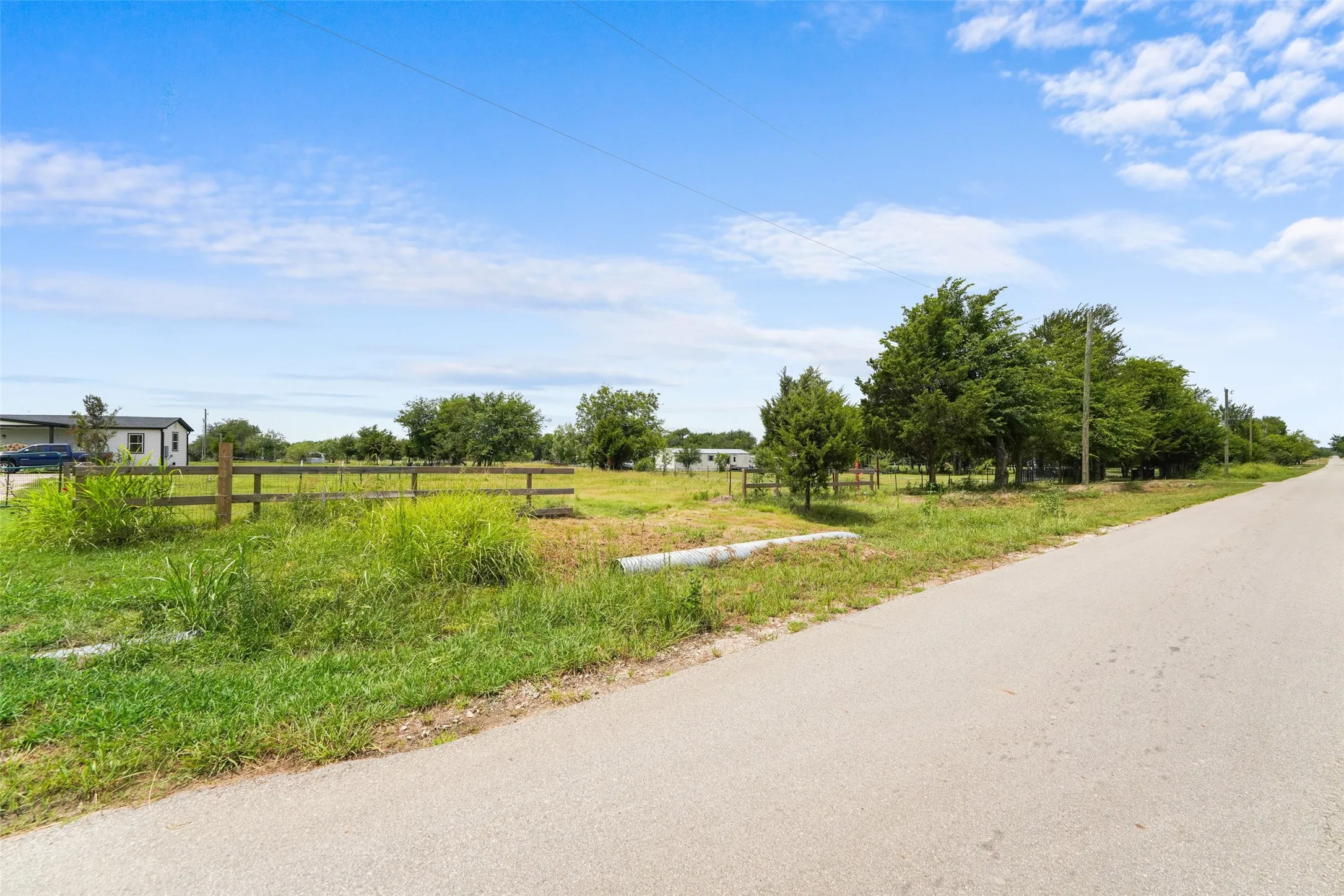 Expansive land parcel featuring a paved roadway, existing fence line, mature trees, and open grassy areas