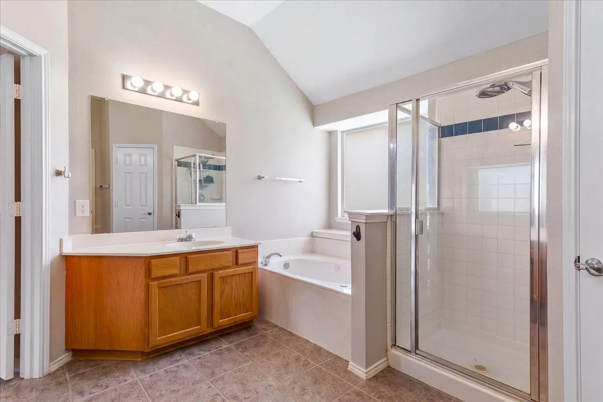 Bathroom featuring a wood-finish vanity with a single sink, a separate glass-enclosed shower, a built-in soaking tub, tile flooring, and a vaulted ceiling