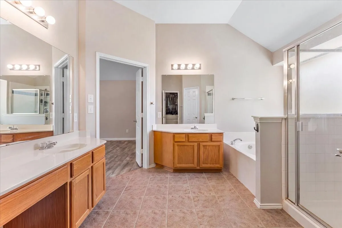 Spacious bathroom featuring twin vanities with integrated sinks and wood cabinetry, a tiled shower with a glass enclosure, an integrated soaking tub, and vaulted ceilings
