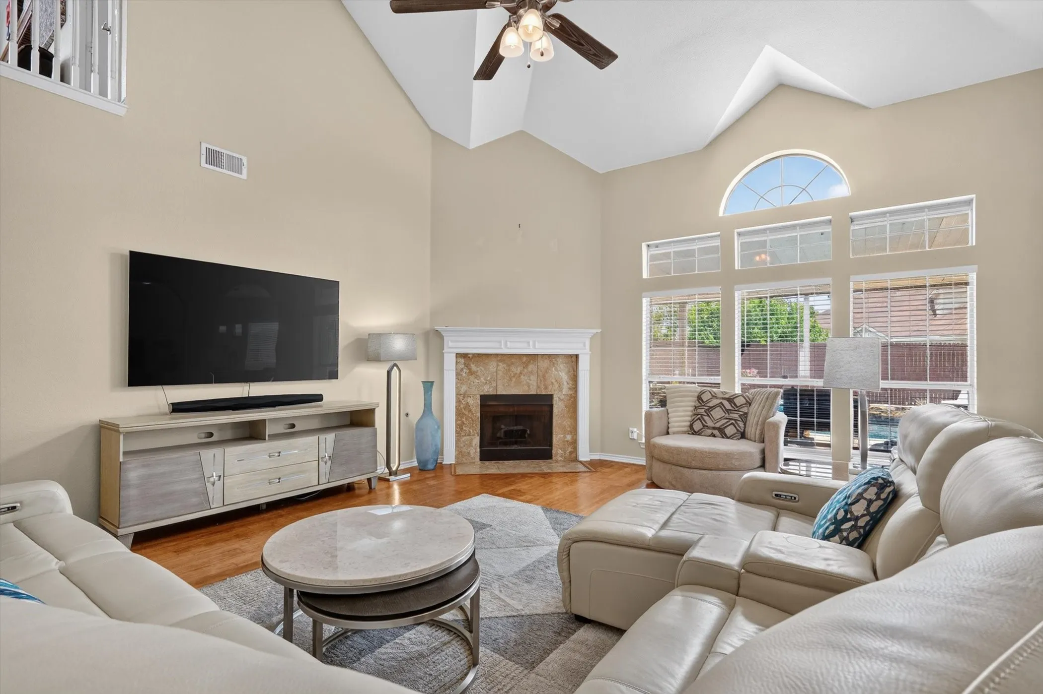 Living area featuring a vaulted ceiling with a ceiling fan, a fireplace with a decorative mantel and stone-finish surround, hardwood-finish flooring, and multiple windows including an arched transom window