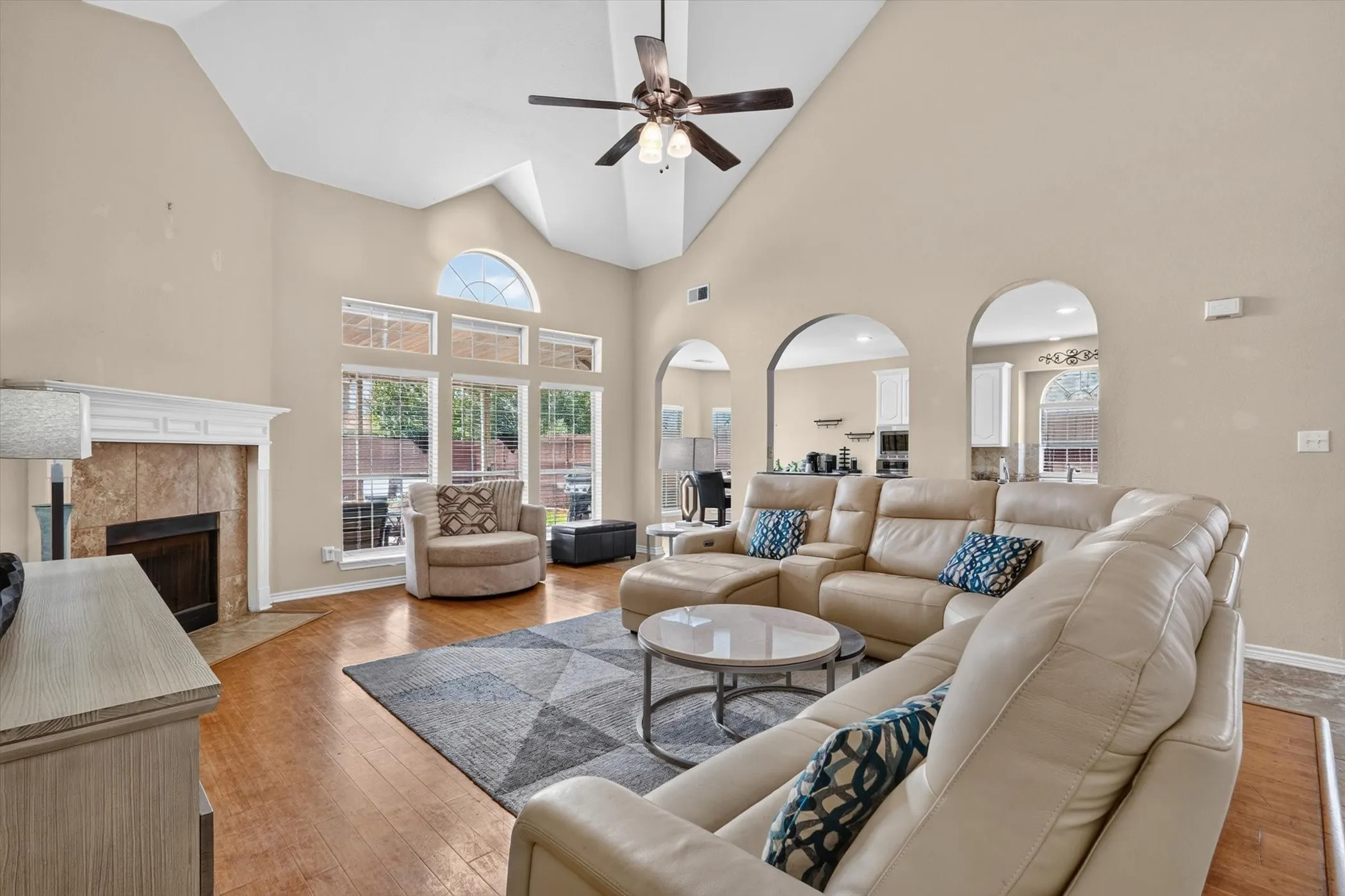 Vaulted ceiling with a ceiling fan, hardwood-finish flooring, fireplace with a tile surround and white mantel, large windows with an arched transom, and arched openings