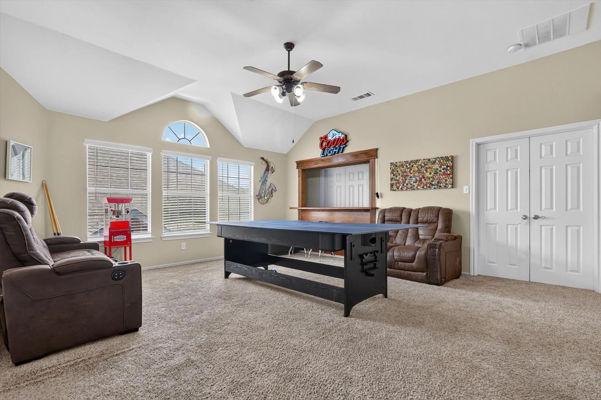 Spacious bonus room featuring vaulted ceilings, a ceiling fan with integrated lighting, three windows with blinds, an arched transom window, and a built-in wood bar