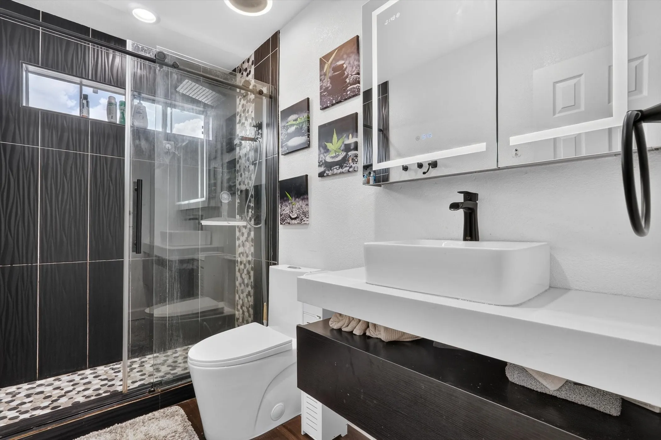 Bathroom featuring a glass-enclosed shower with pebble-style floor tiling, a white vessel sink, a wall-mounted vanity, and an illuminated medicine cabinet