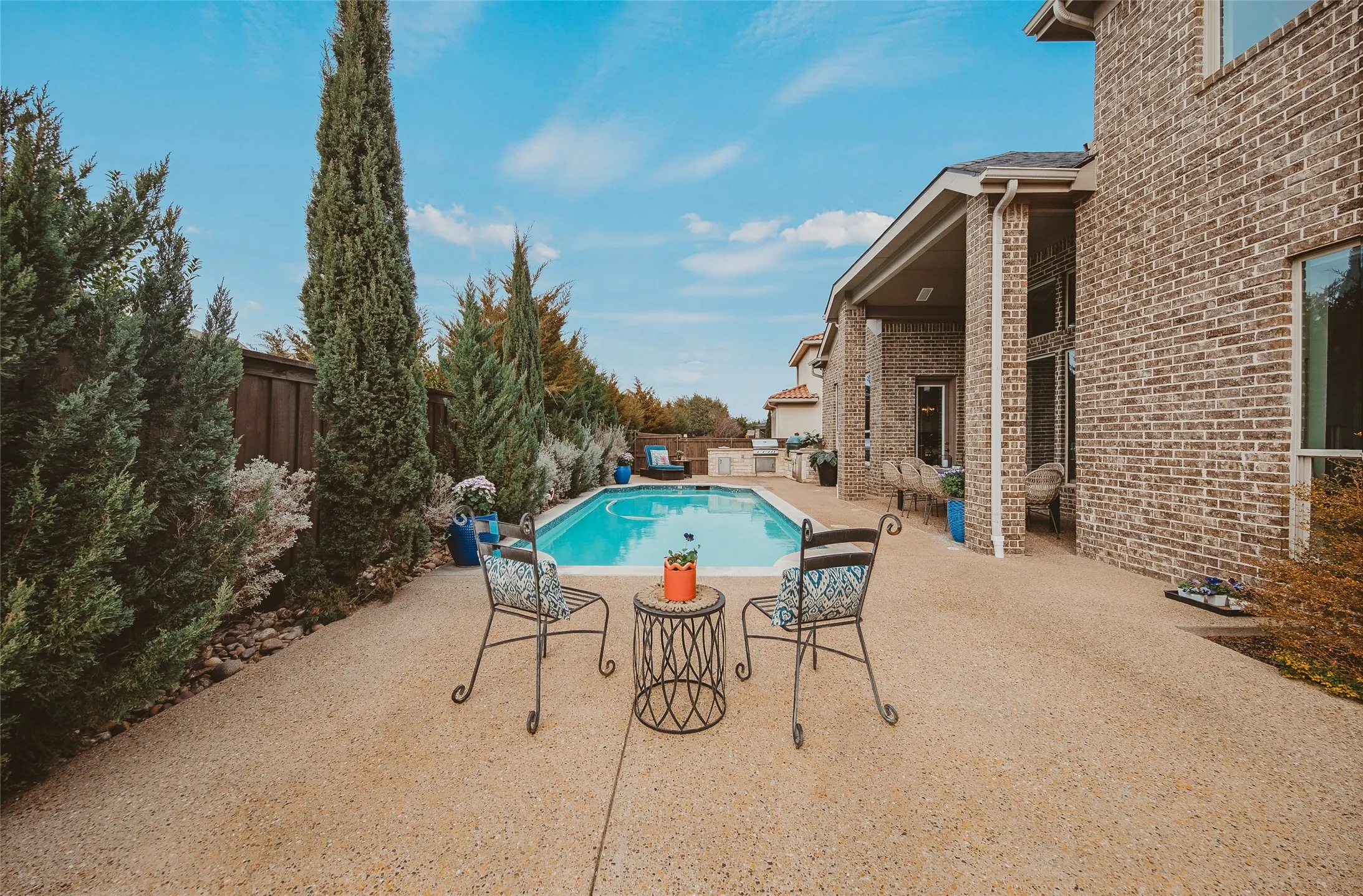 Outdoor pool area featuring a perimeter of mature evergreens, a textured concrete patio, a rectangular swimming pool, and a brick exterior facade