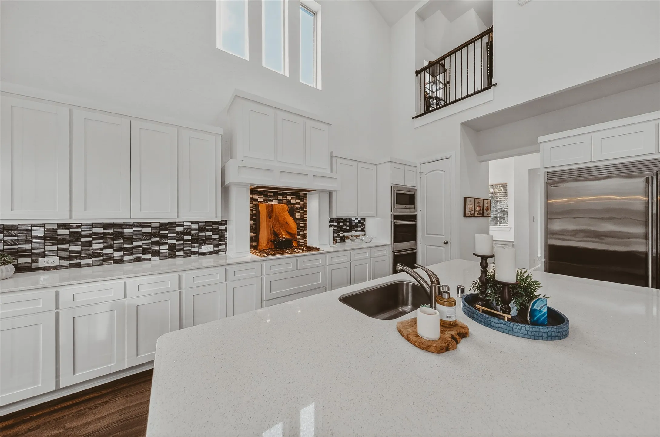 Spacious kitchen featuring an oversized island with an undermount sink, white cabinetry, a mosaic tile backsplash, stainless steel appliances, and wood-finish flooring