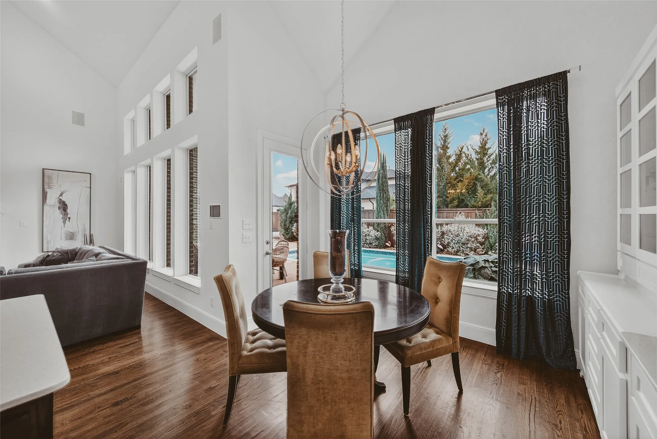 Dining area featuring rich wood-finish flooring, a contemporary chandelier, and large windows with patterned privacy curtains