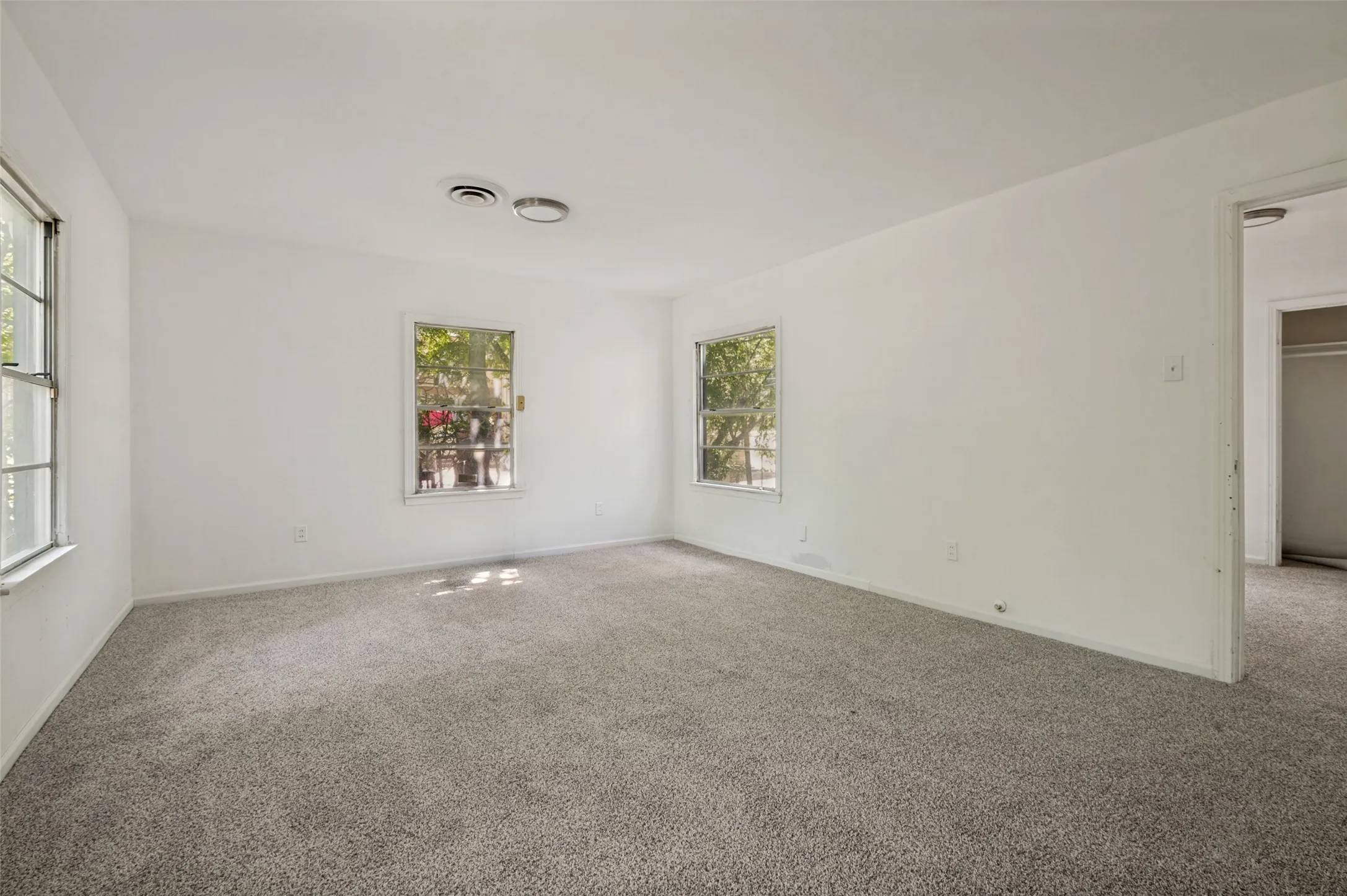 Spacious interior room featuring light gray carpeting, neutral white walls, two casement windows with exterior foliage, and ceiling-mounted vents