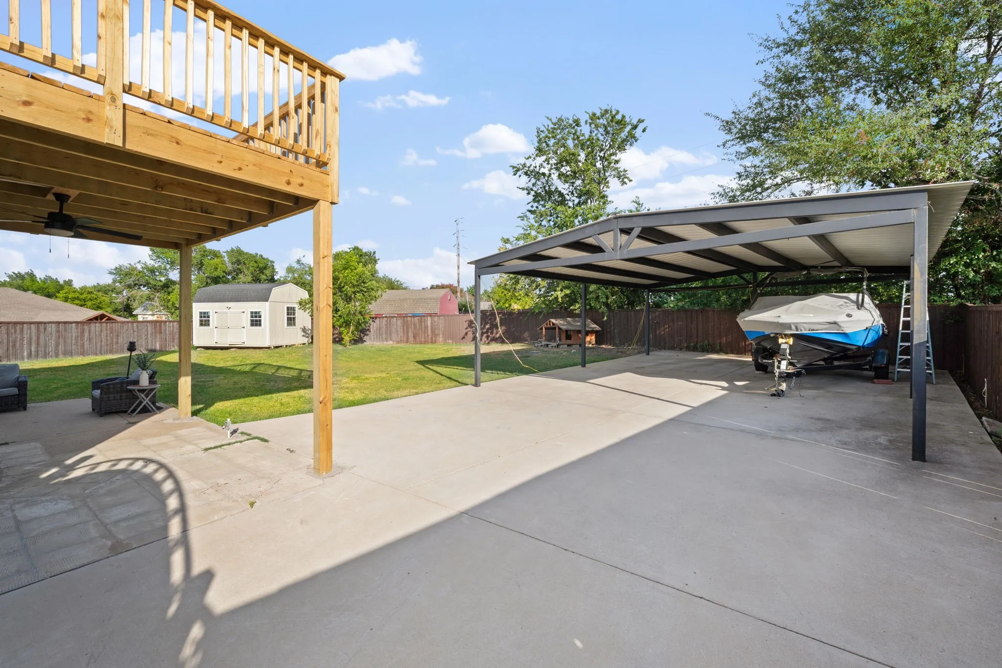 Expansive concrete patio area featuring a covered carport, an elevated wood deck, a ceiling fan, and a detached storage shed on a grassy lawn