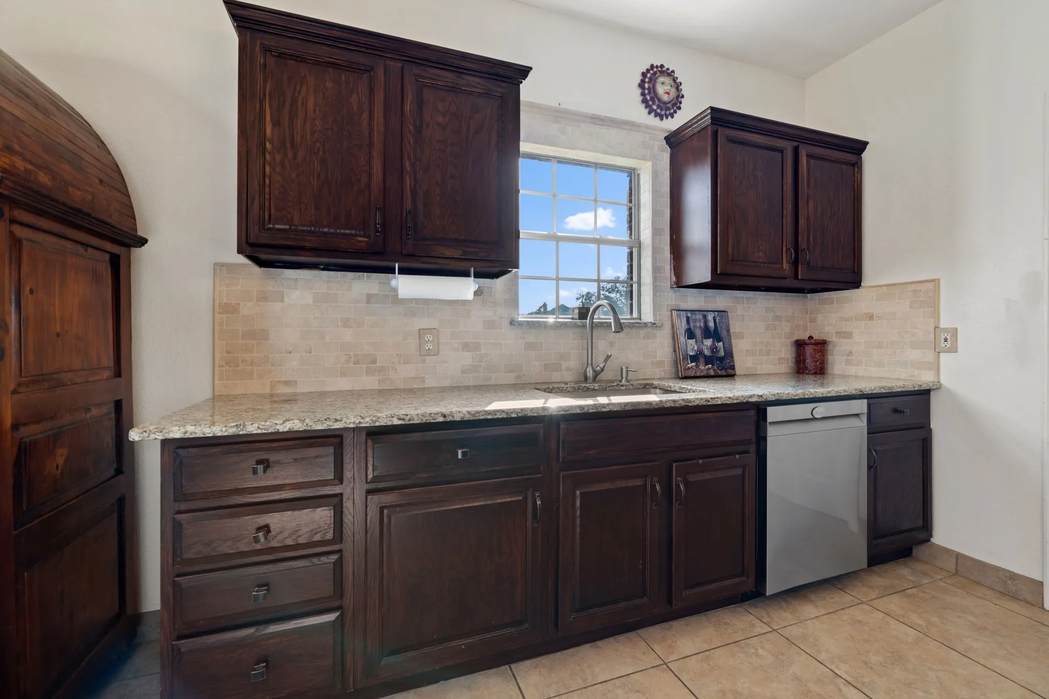 Kitchen featuring dark wood cabinetry, light-toned tiled flooring, a stainless steel sink with a single-handle faucet, a light granite countertop, and a tiled backsplash
