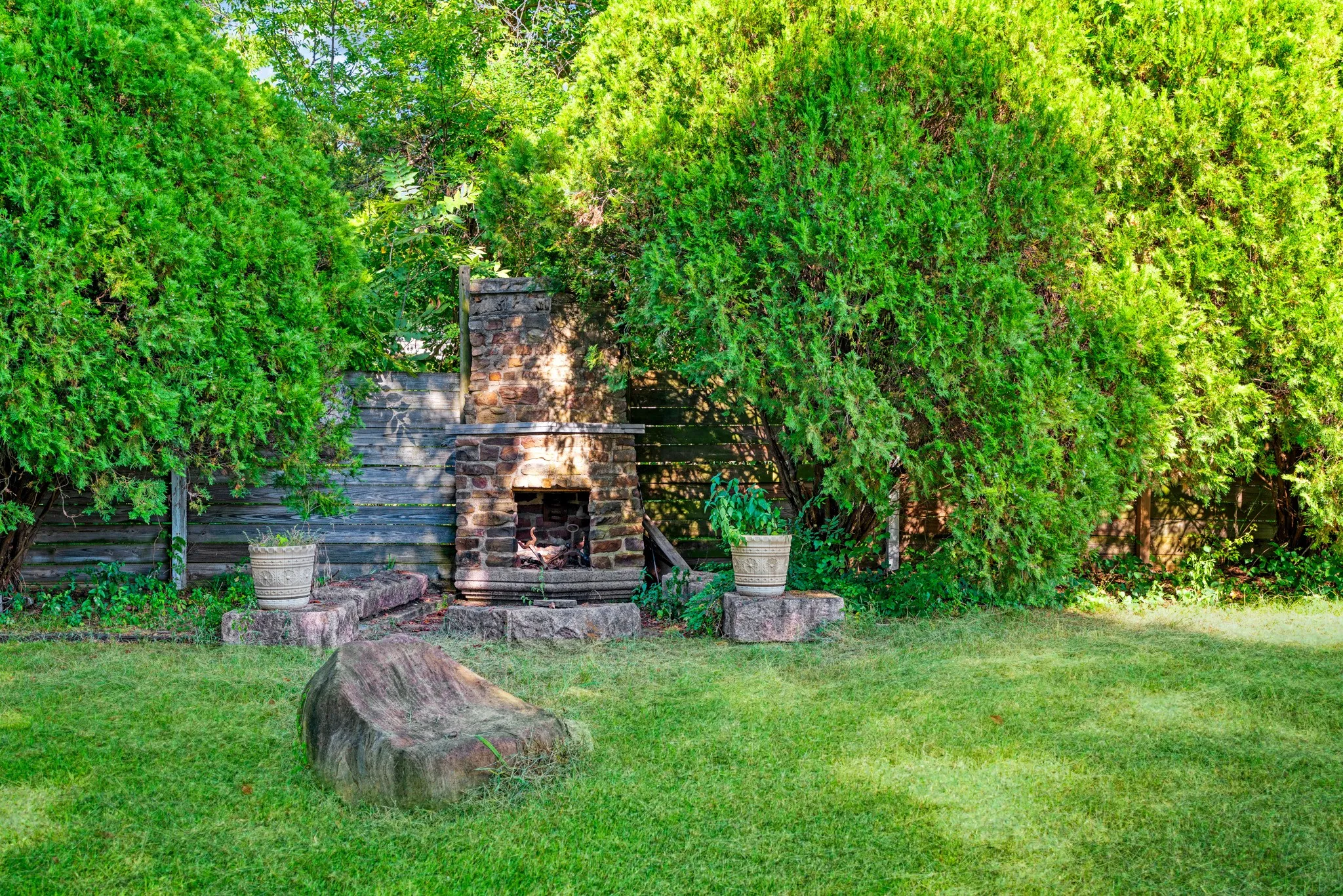 Expansive lawn area featuring a stone fireplace with a hearth, bordered by dense green foliage