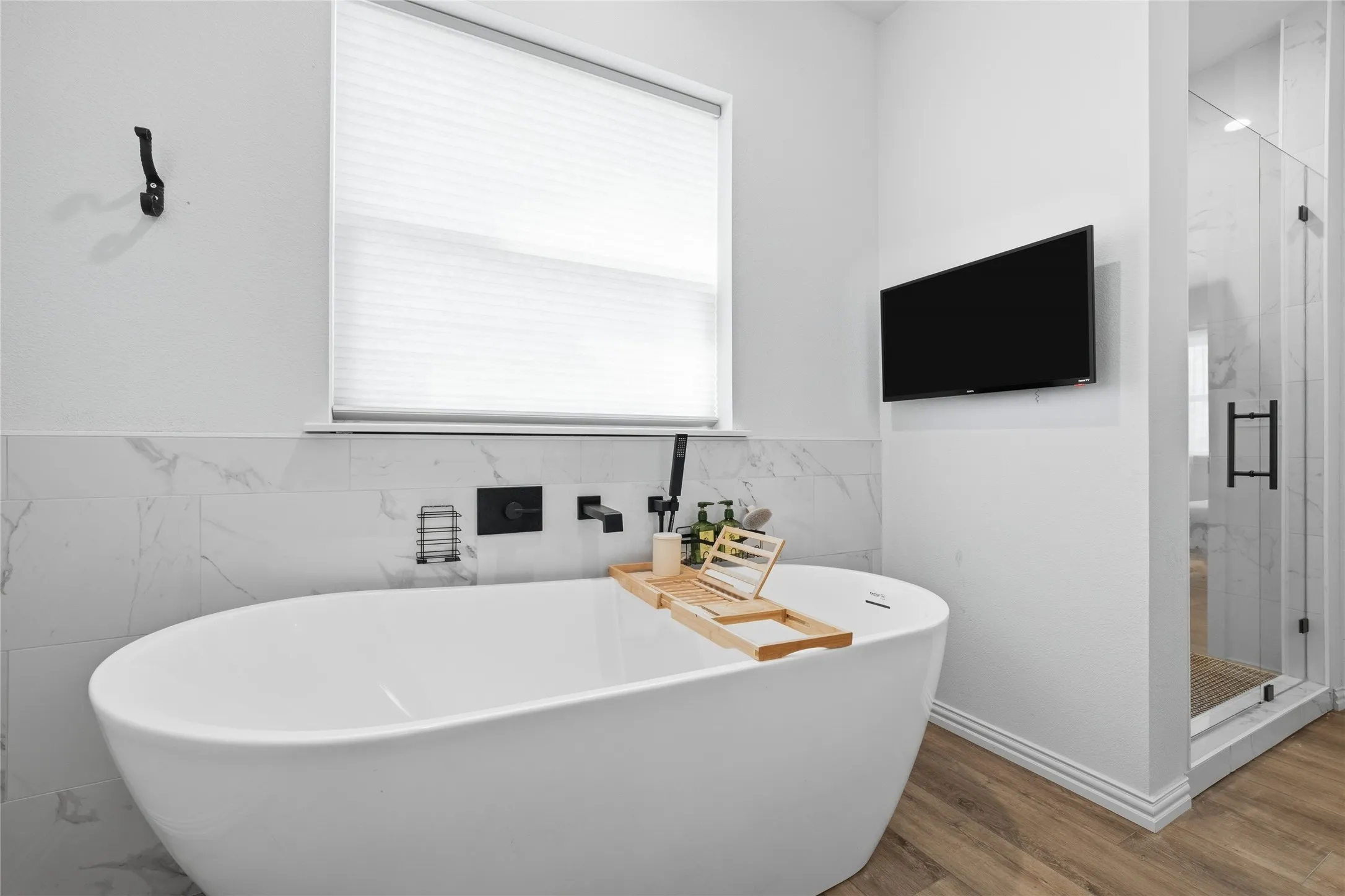 Bathroom featuring a freestanding oval tub, wall-mounted matte black fixtures, marbled tile wainscoting, and wood-finish flooring