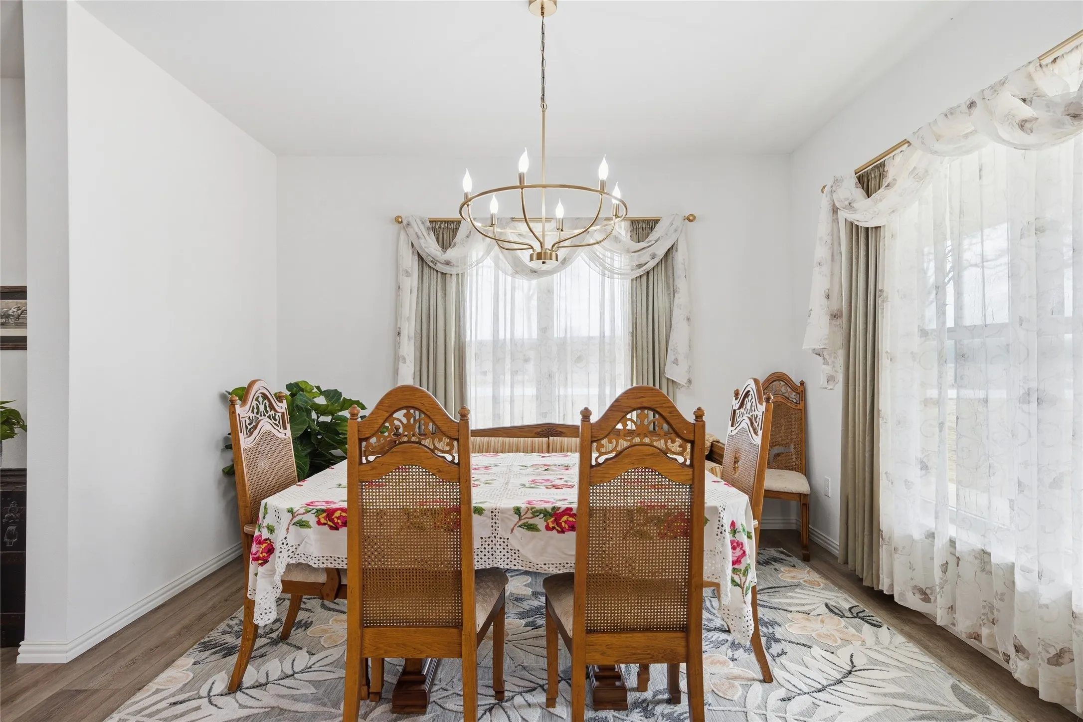 Dining area featuring wood-finish flooring, a contemporary chandelier, and large windows with sheer and opaque drapery