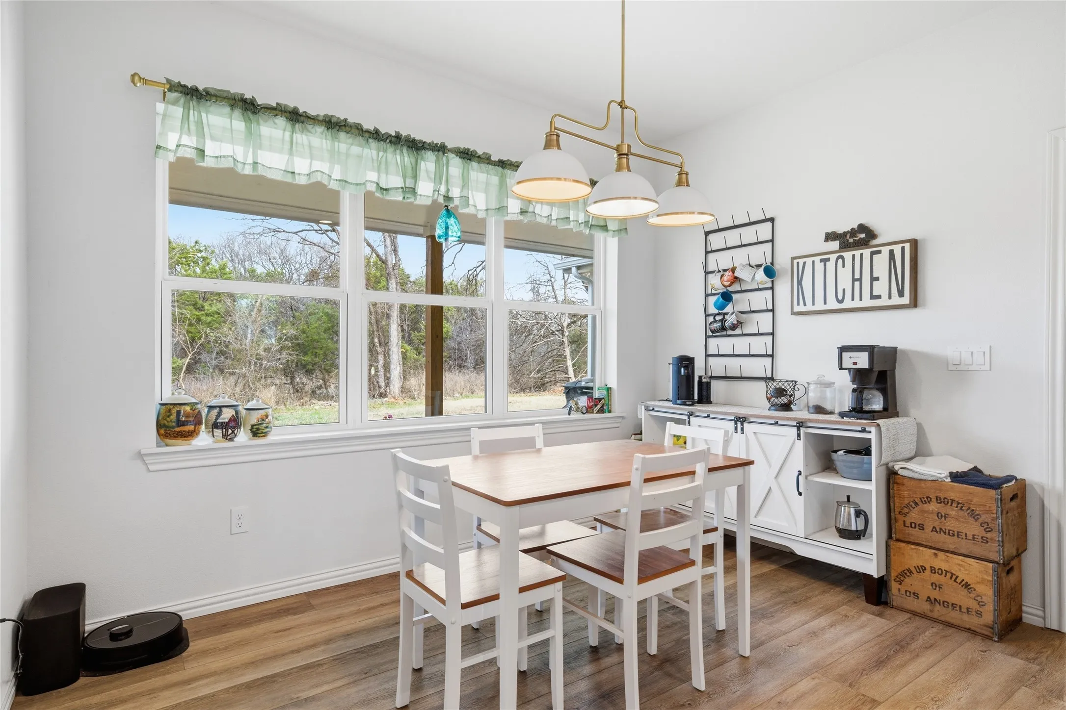 Bright kitchen workspace featuring a wood-finish dining table with seating for four, white cabinetry, stainless steel appliances, and abundant natural light from large multi-pane windows overlooking the landscape