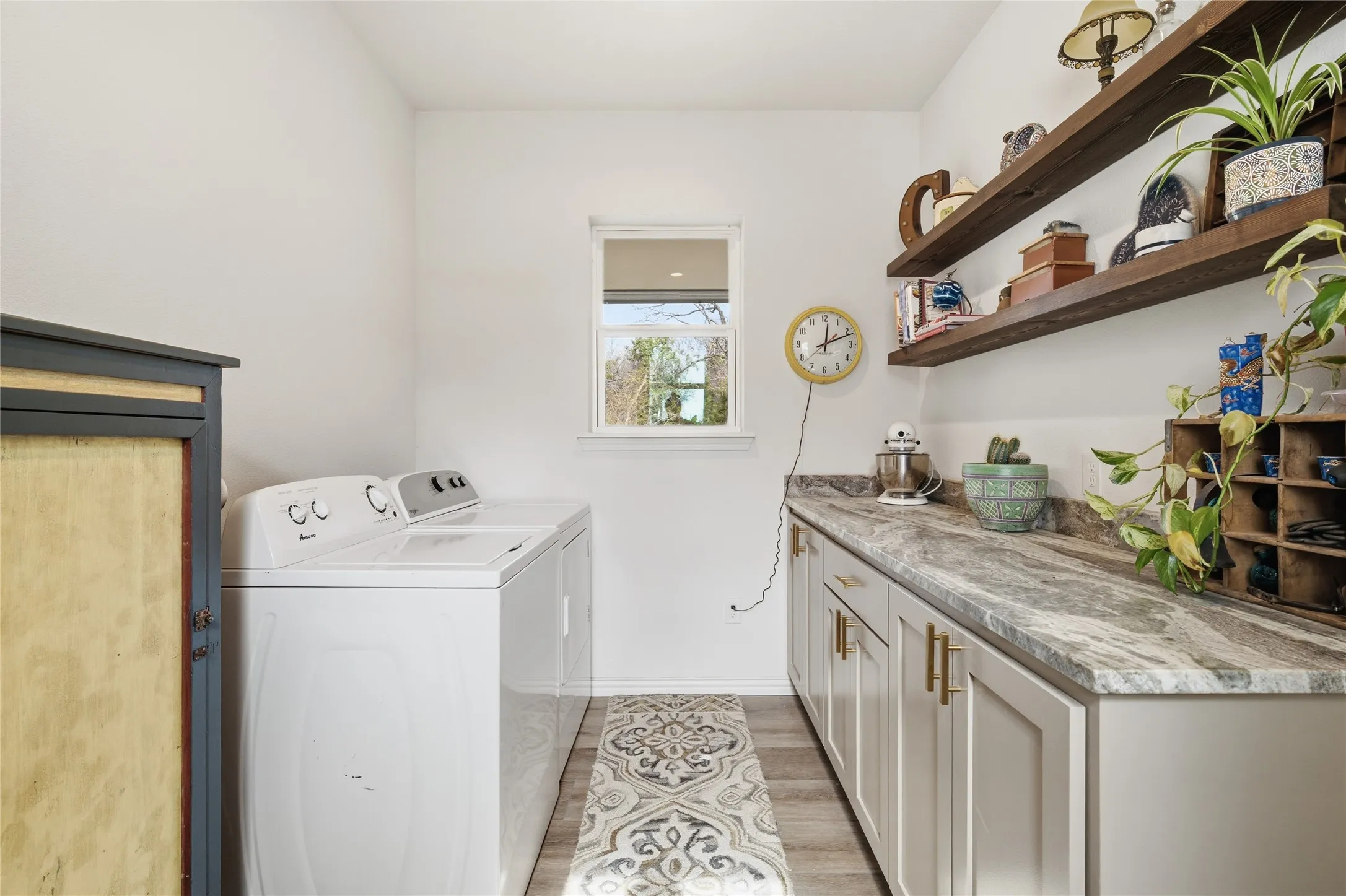 Dedicated laundry room featuring a top-load washer with intuitive controls, white cabinetry with wood-finish countertops, open shelving displaying storage solutions, a small window for natural light, wall-mounted clock, and patterned runner flooring for functionality and style