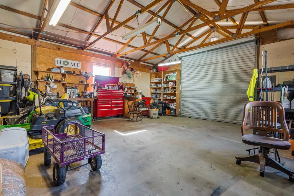 Spacious workshop featuring exposed wood trusses, concrete flooring, built-in shelving, a large roll-up door, and fluorescent lighting