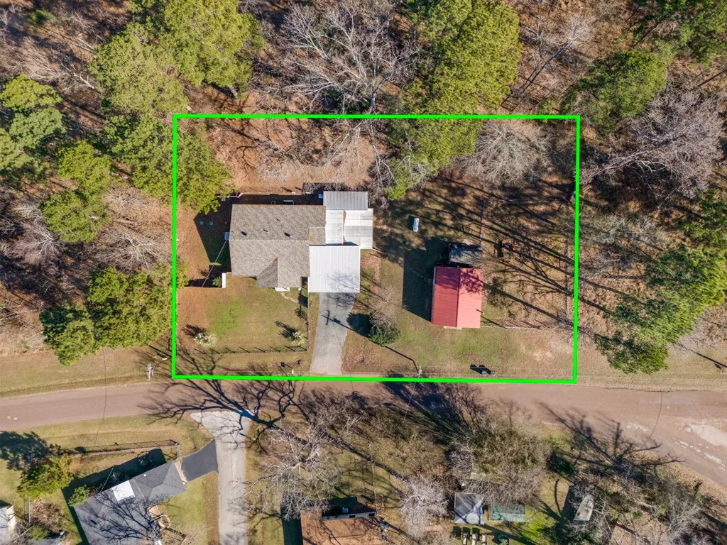 Aerial perspective showcasing a property surrounded by mature trees, featuring a main residential structure with a gray roof, a separate red-roofed outbuilding, and a paved driveway leading to the residence