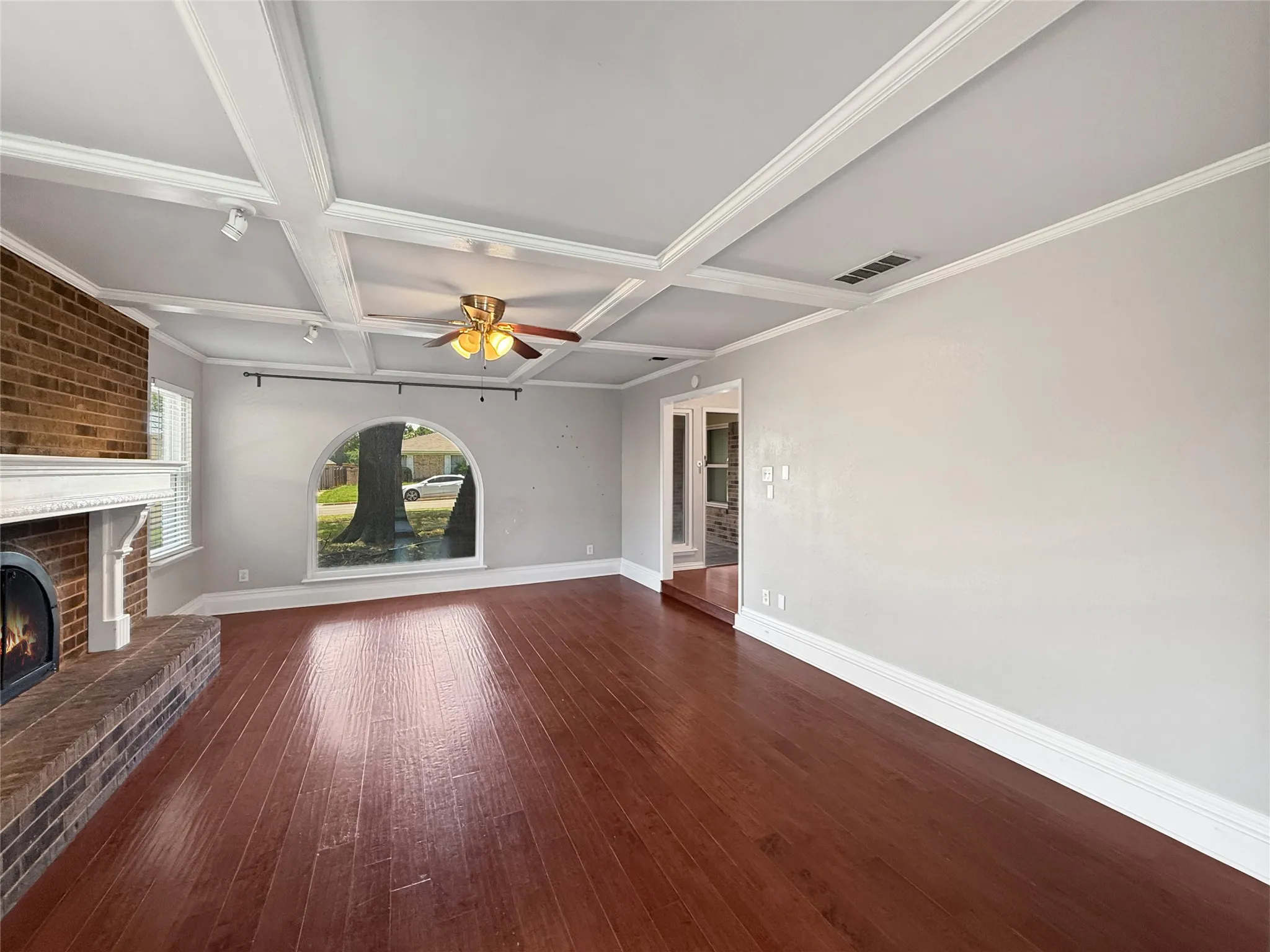 Spacious living area featuring wood-finish flooring, a brick fireplace with a white mantel, an arched window, a coffered ceiling with a ceiling fan, and track lighting