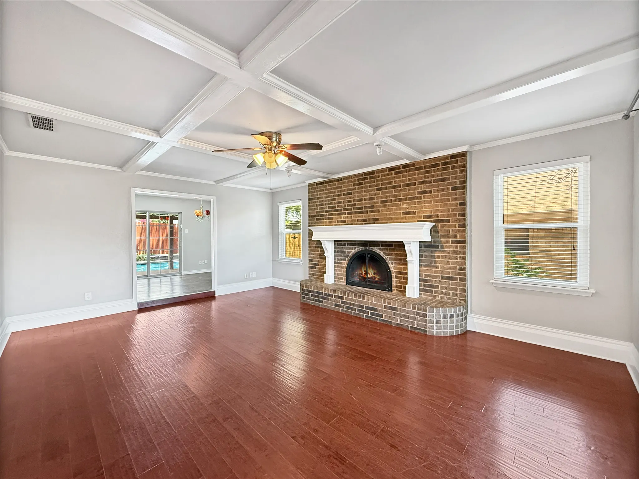 Spacious room featuring wood-finish flooring, a coffered ceiling with a ceiling fan, and a brick fireplace with a white mantel