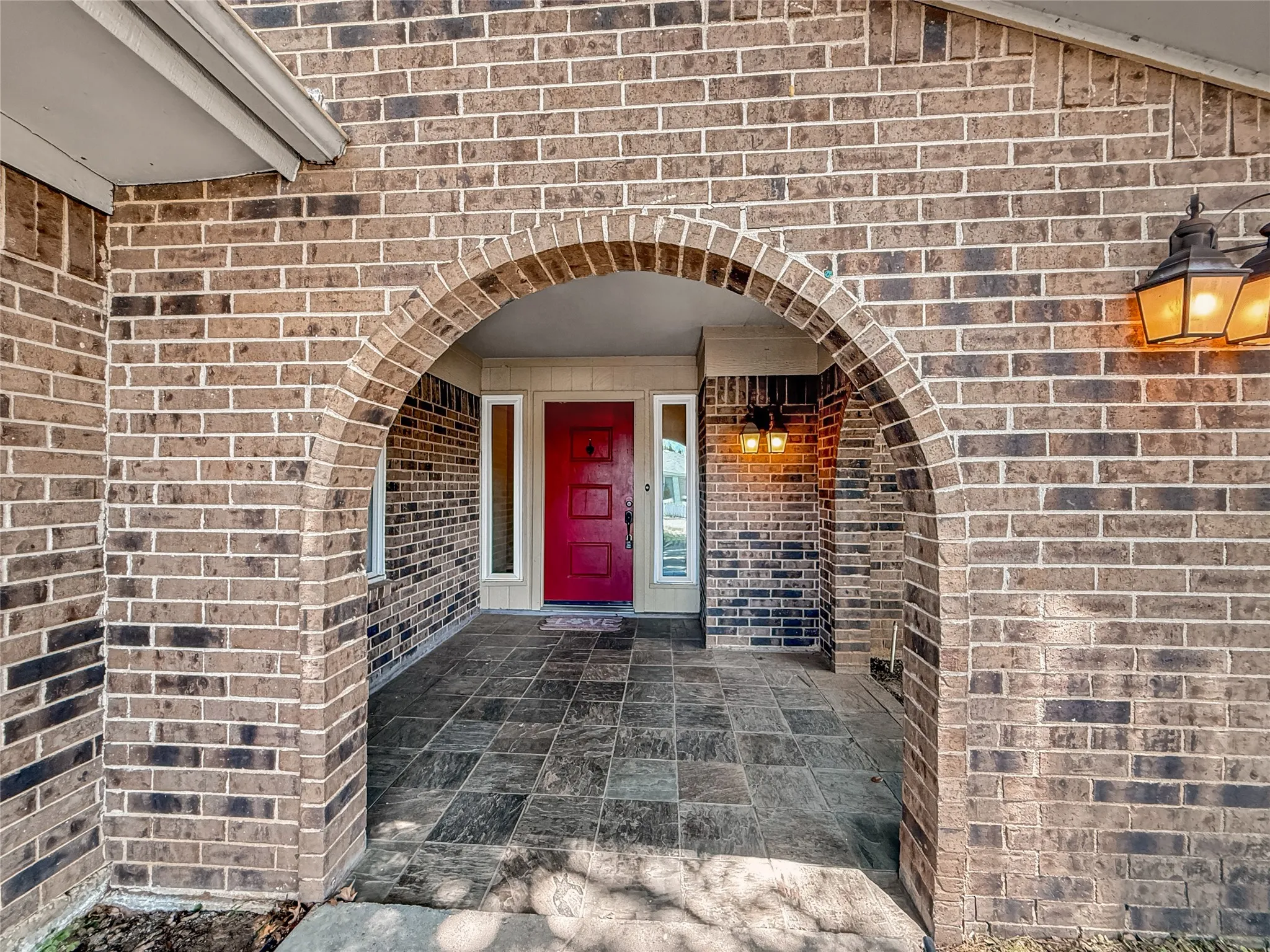 Brick exterior featuring an arched entry, a red front door, transom windows, exterior sconces, and slate-style flooring
