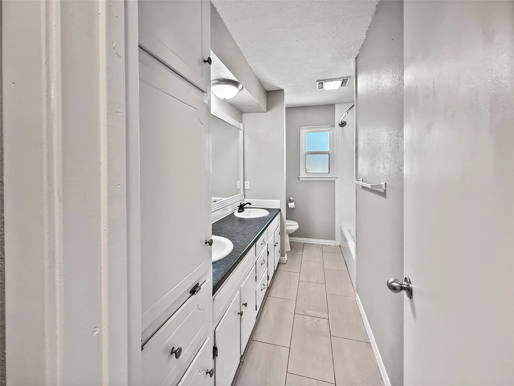 Bathroom featuring a dual vanity with undermount sinks, a dark countertop, white cabinetry with various storage options, and light-toned plank tile flooring