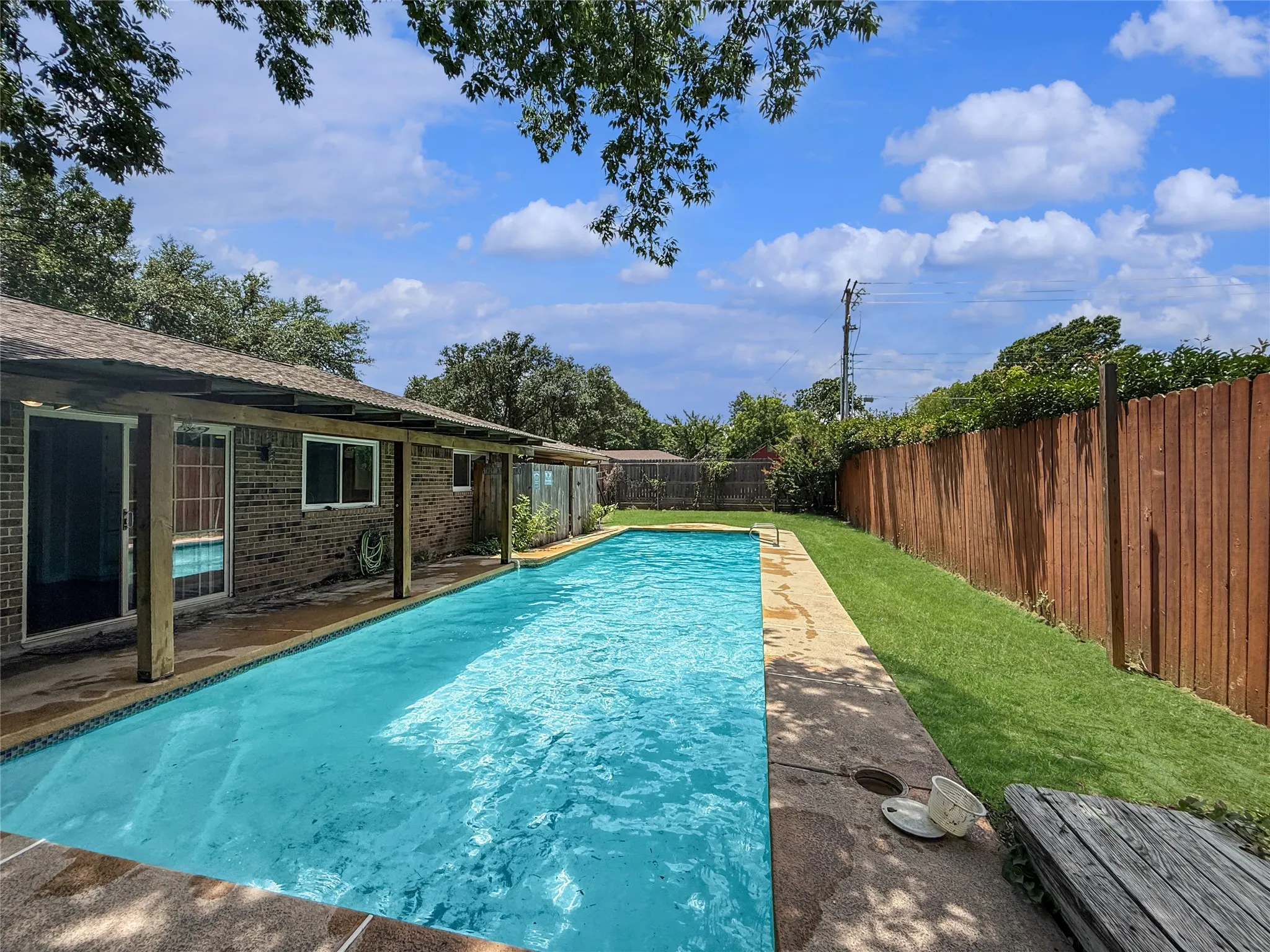 Expansive backyard featuring a rectangular swimming pool with concrete decking, a brick exterior, a covered patio, and a wood privacy fence