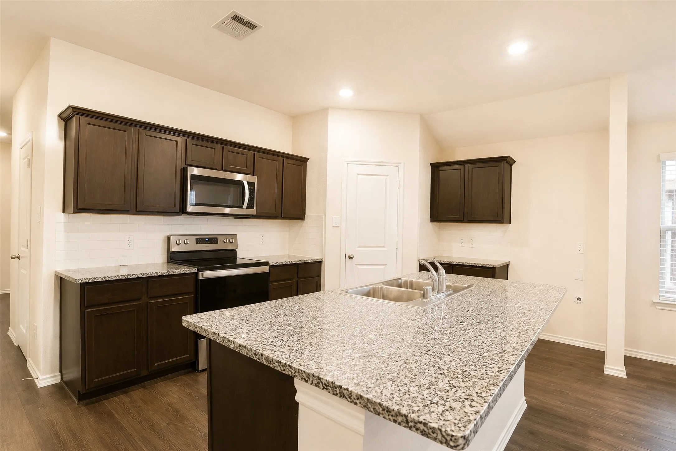 Contemporary kitchen featuring an island with a double basin sink, dark wood-finish cabinetry, light-toned patterned countertops, stainless steel appliances, and wood-finish flooring